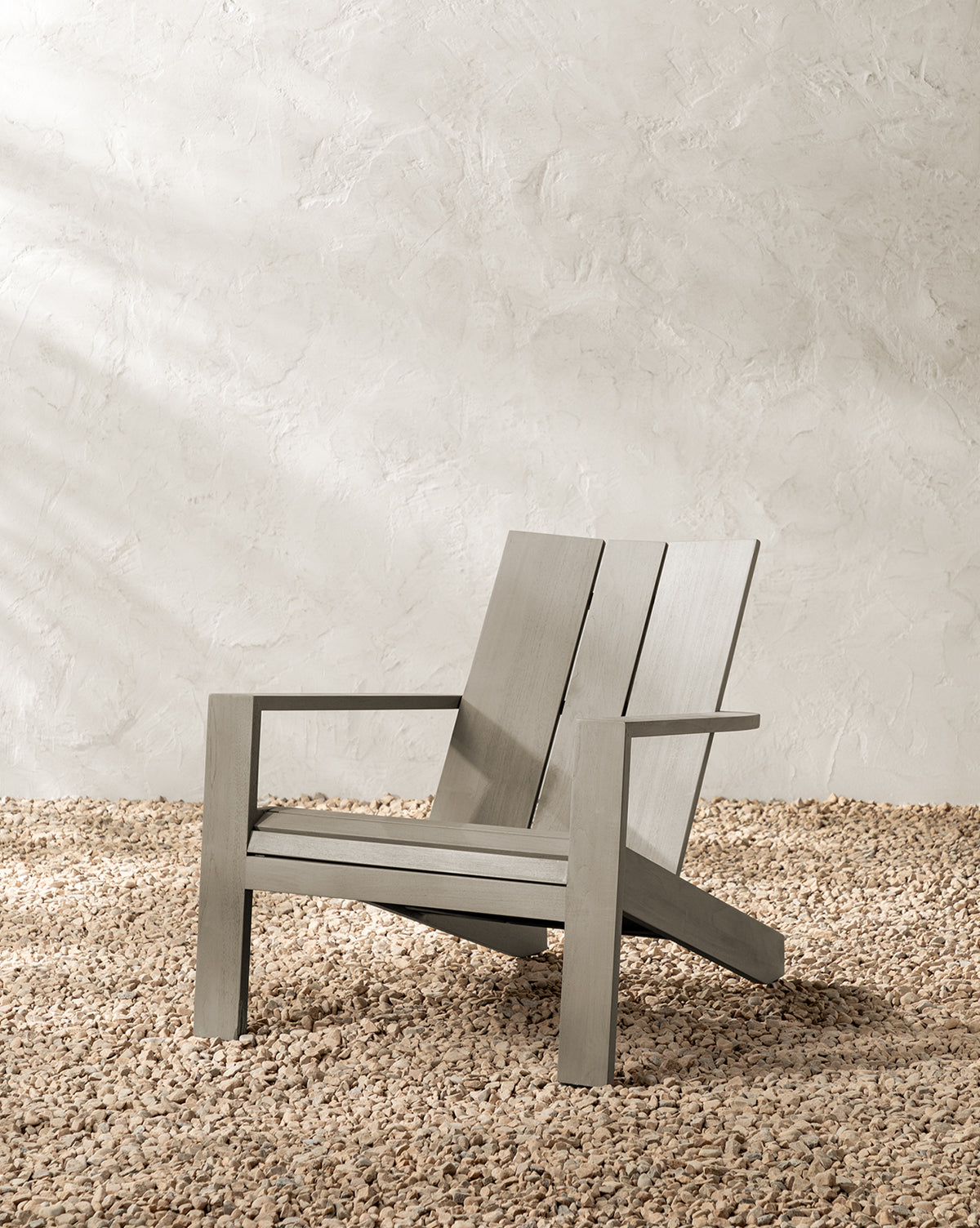 The Triby Gray Teak Outdoor Lounge Chair, featuring wide slats and an angled backrest in a modern minimalist design, rests on light-colored pebbles before a textured, off-white wall, by McGee & Co.
