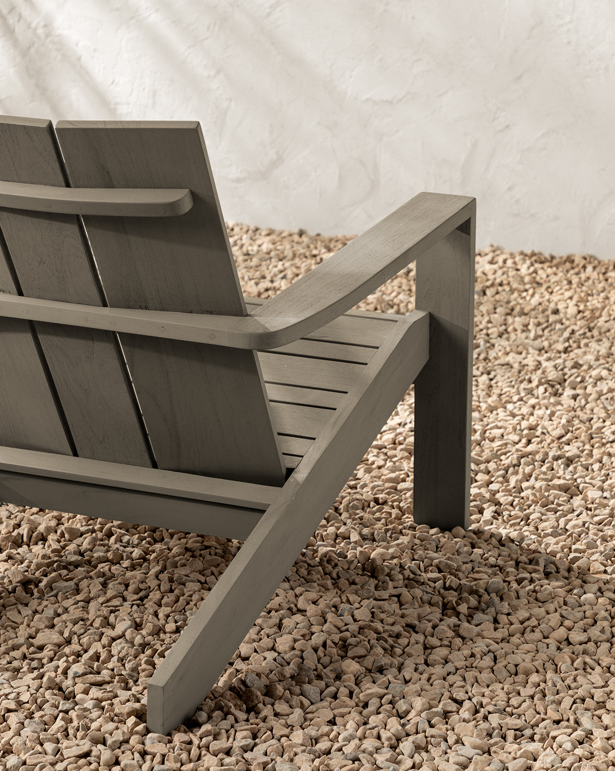 A close-up of the back and armrest of the Triby Gray Teak Outdoor Lounge Chair on light-colored gravel, set against a textured white wall, by McGee & Co.