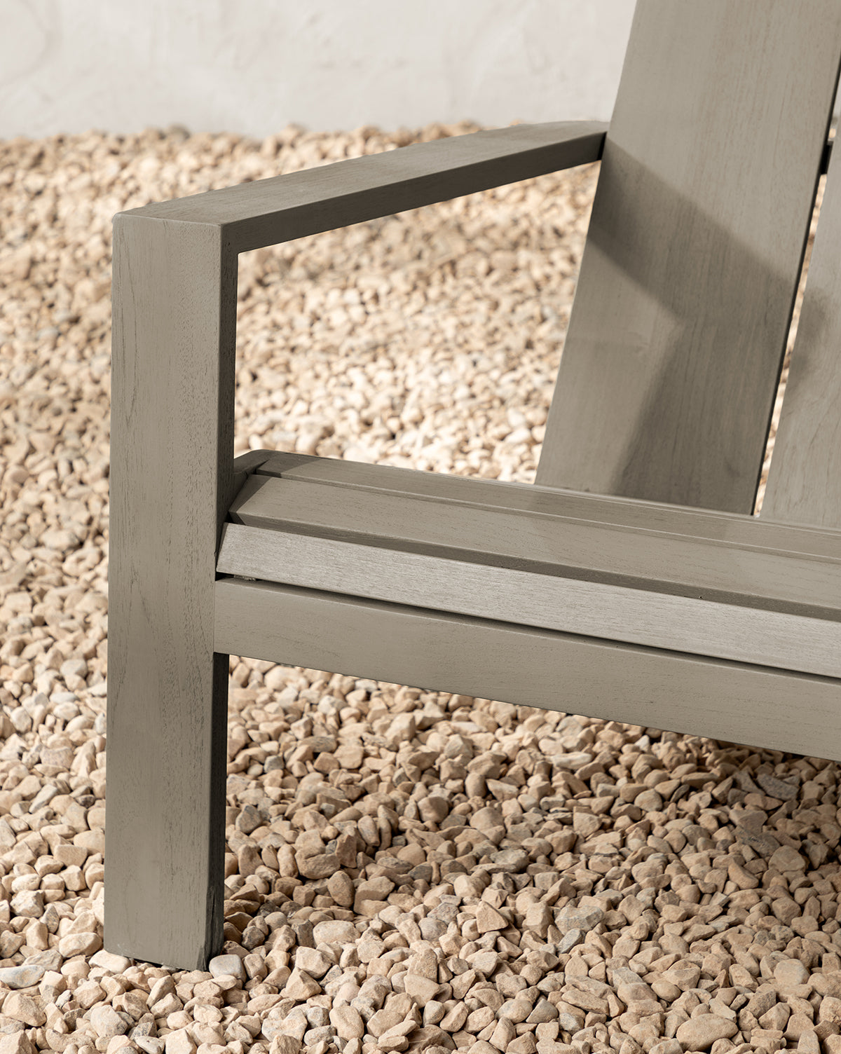 Close-up of the Triby Gray Teak Outdoor Lounge Chair, featuring modern angular armrests, clean lines, and a minimalist design, by McGee & Co. The light gray teak chair is shown on a bed of small, light-colored pebbles.