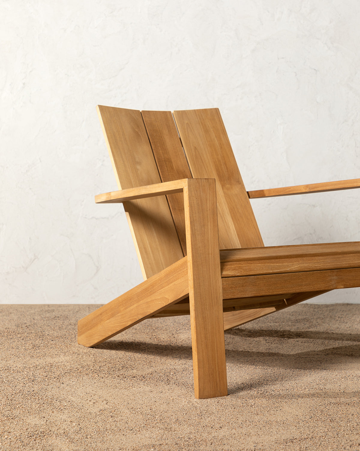 The Triby Teak Outdoor Lounge Chair, featuring wide angled slats and armrests, sits on a textured sandy patio floor against a light-colored wall by McGee & Co.