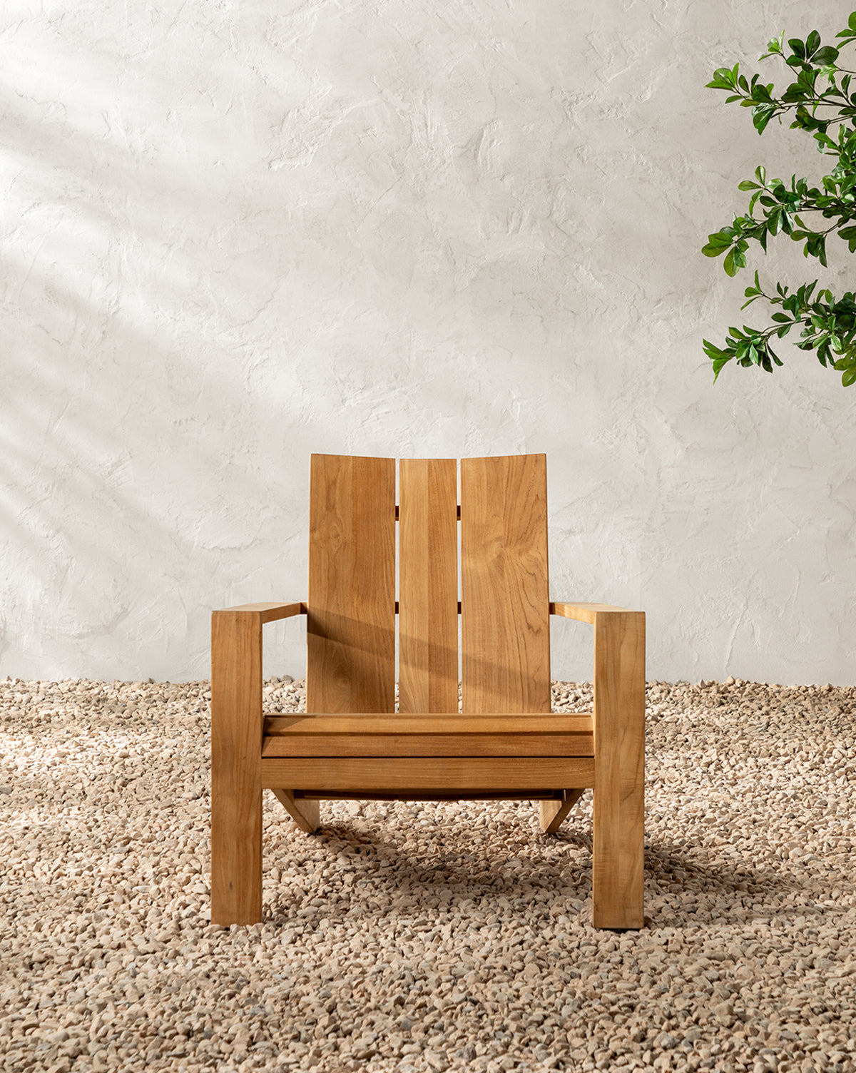 The Triby Teak Outdoor Lounge Chair rests on pale pebbles by a textured white wall, while sunlight creates gentle shadows and a leafy green plant peeks in from the right, highlighting this tranquil, all-weather setting, by McGee & Co.