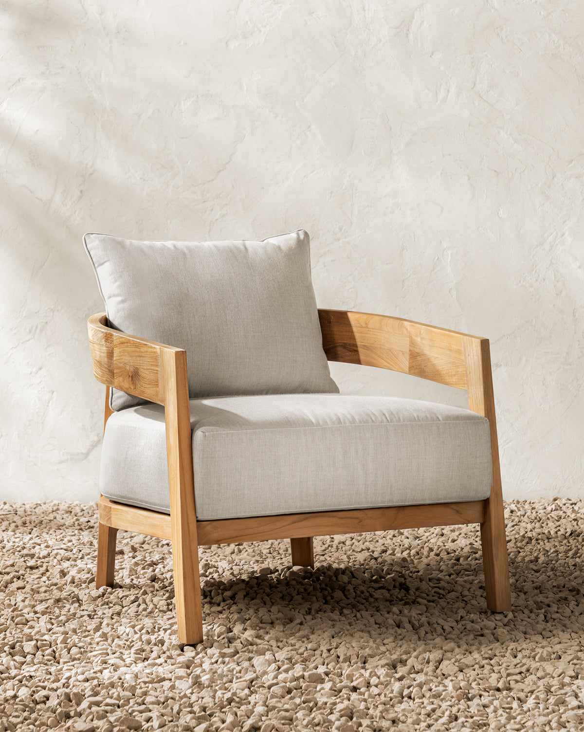 The Vento Teak Outdoor Lounge Chair, featuring a light wood frame and pale beige cushions, rests on a textured stone floor against a cream-colored wall, by McGee & Co.