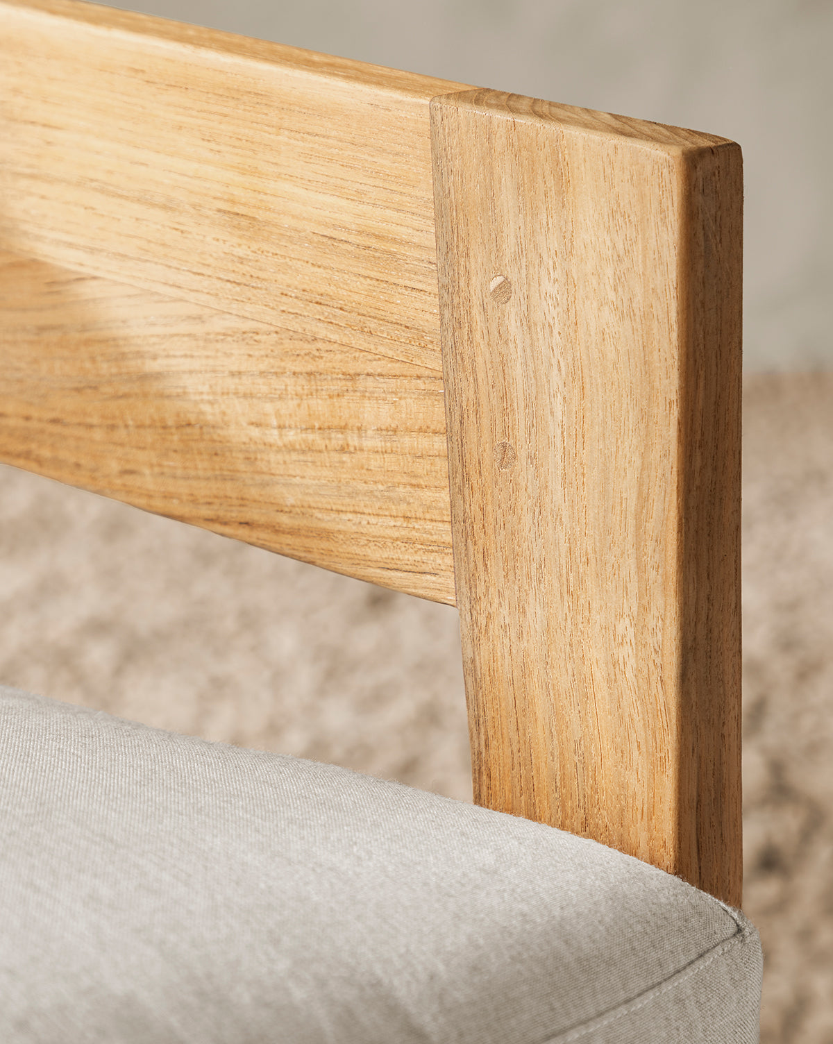 Close-up of the Vento Teak Outdoor Lounge Chairs light teak wood backrest and frame, featuring a light beige cushioned seat with a blurred beige carpet in the background, by McGee & Co.