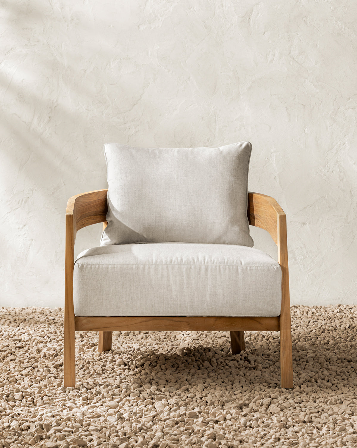 The Vento Teak Outdoor Lounge Chair, featuring a light wood frame and light beige cushions, sits on a textured beige carpet against a light-colored, textured wall, by McGee & Co.