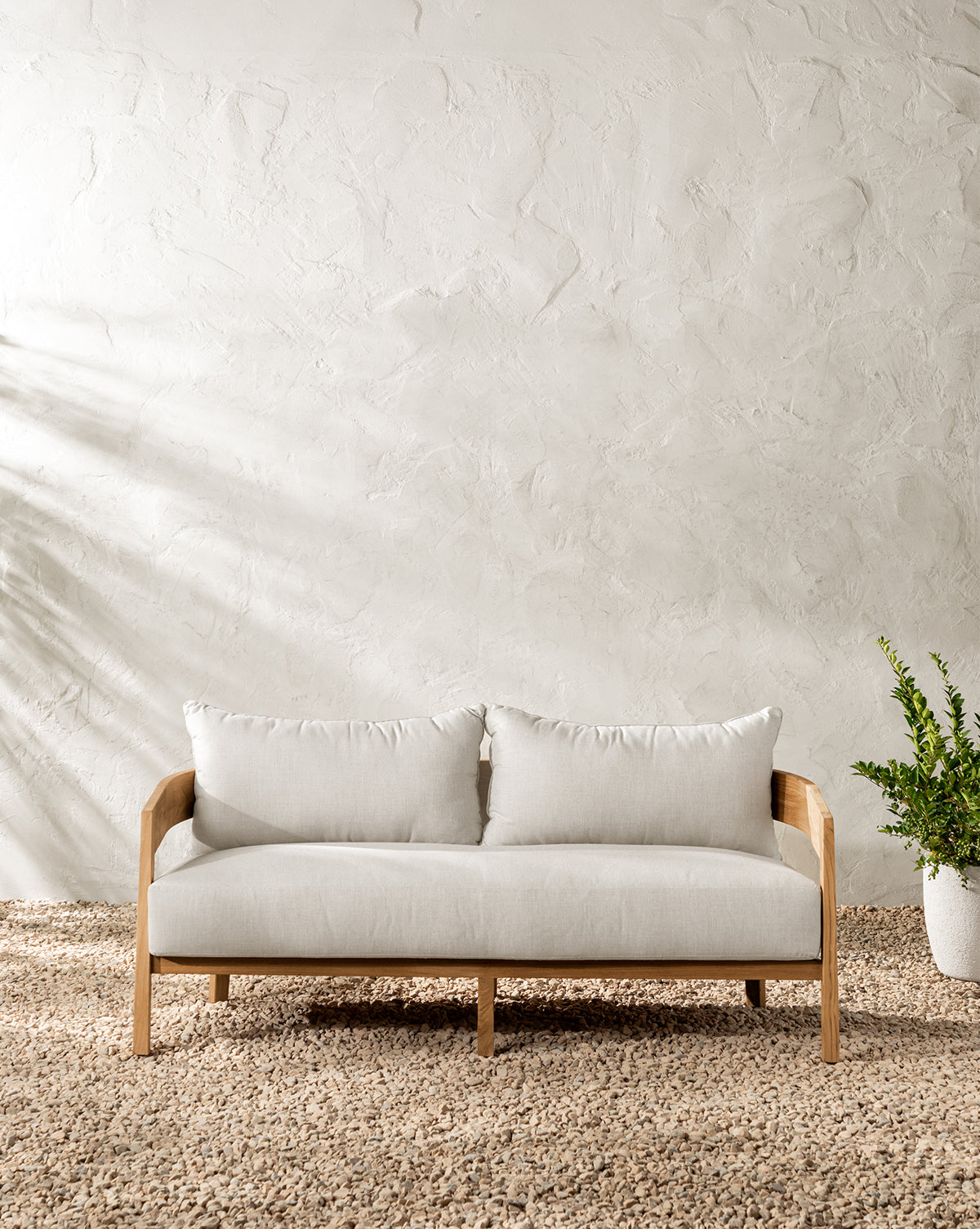 The Vento Outdoor Sofa 68.75 with light Sunbrella cushions sits on a textured beige floor by a white stucco wall. A green potted plant stands to the right, while sunlight casts soft shadows across this stylish outdoor space by McGee & Co.