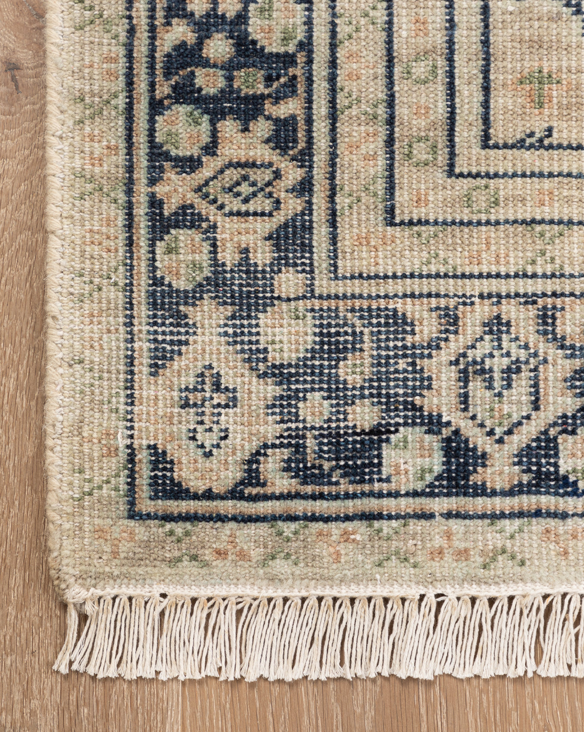 A close-up of the Vercelli Hand-Knotted Wool Rug corner shows its fringed edges and geometric floral patterns in navy blue, beige, and green on a light wooden floor, by McGee & Co.
