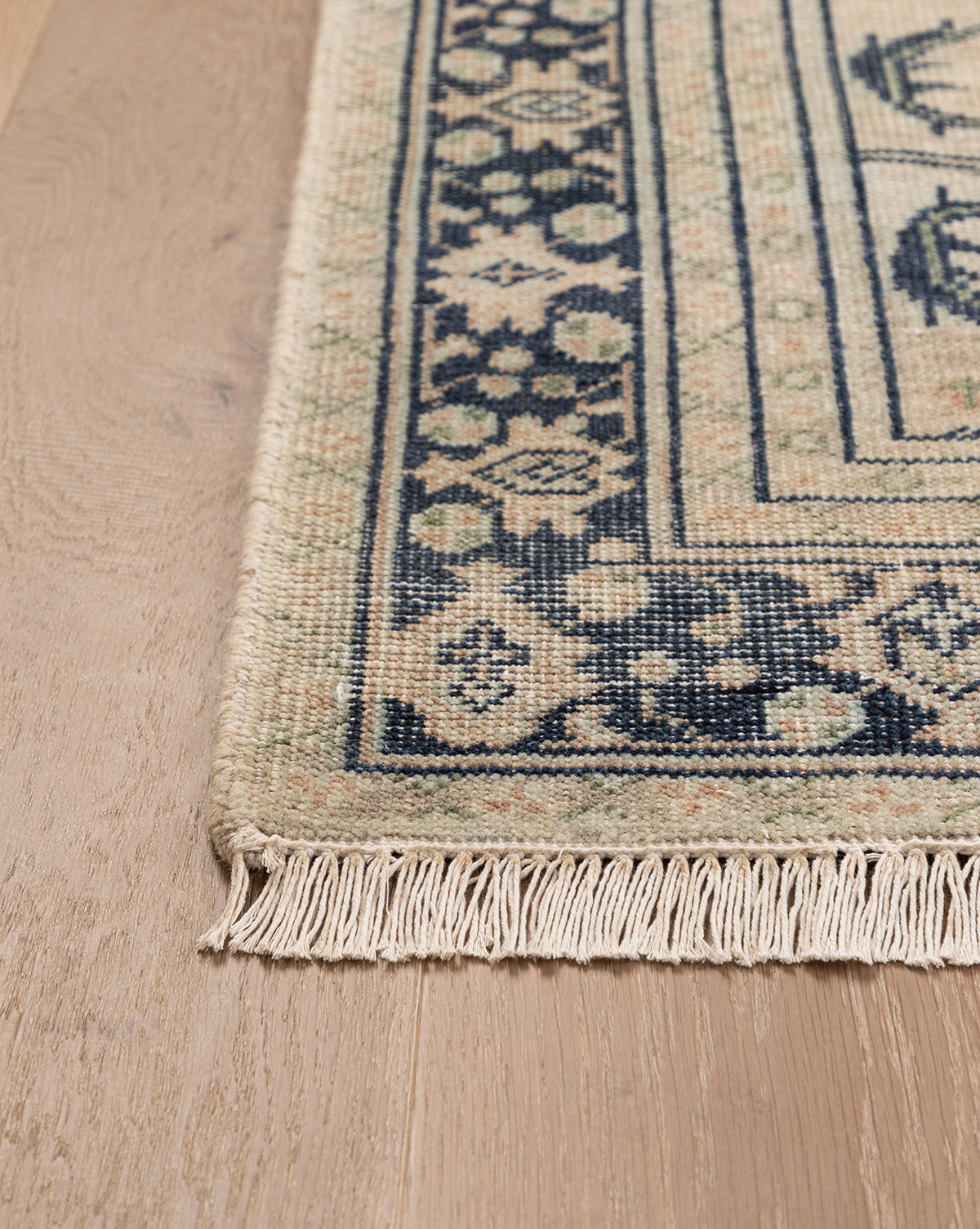 A close-up of the Vercelli Hand-Knotted Wool Rug with fringe, shown on a light wood floor. The rug displays beige and navy geometric and floral border motifs, by McGee & Co.