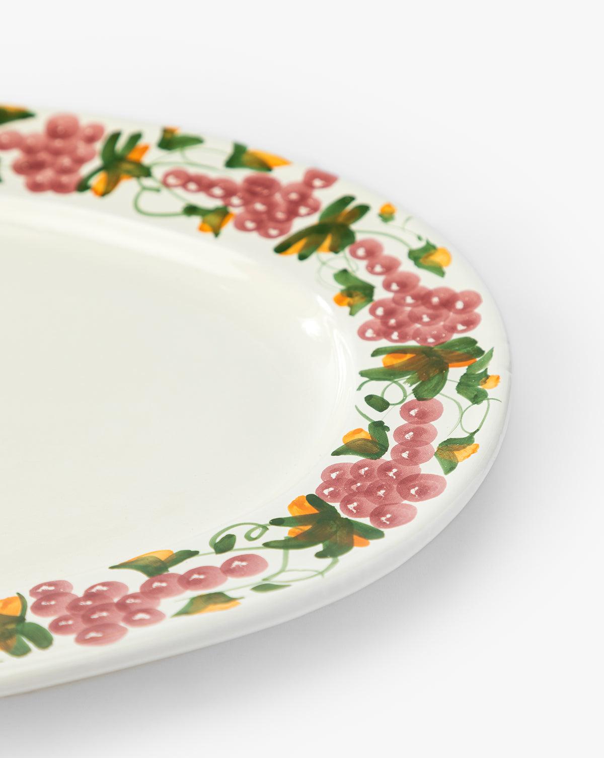 The Vine Serving Platter features a close-up of a white ceramic plate with a decorative grapevine border of painted pink grapes, green leaves, and yellow accents, by McGee & Co.