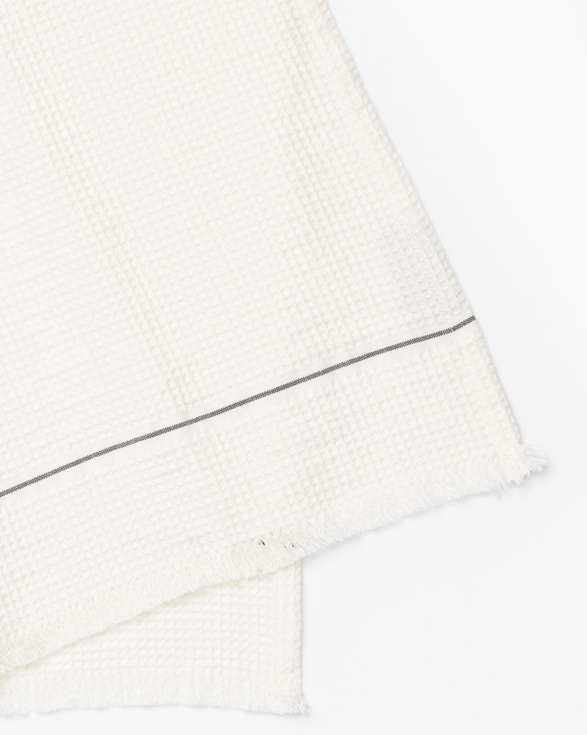 A close-up of the Waffle Hand Towel, made from a linen-cotton blend in a waffle texture with a thin dark stripe and frayed edges, shown on a white background—ideal for adding style to your kitchen decor, by McGee & Co.