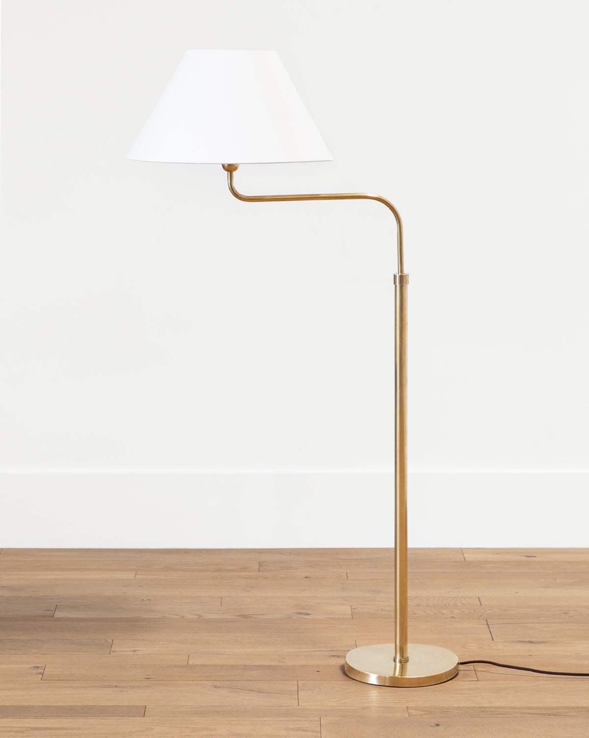 The Wainwright Swoop Floor Lamp features a slim gold metal base and stem, a white angled lampshade, and a circular base, standing elegantly on a wooden floor against a plain white wall, by McGee & Co.