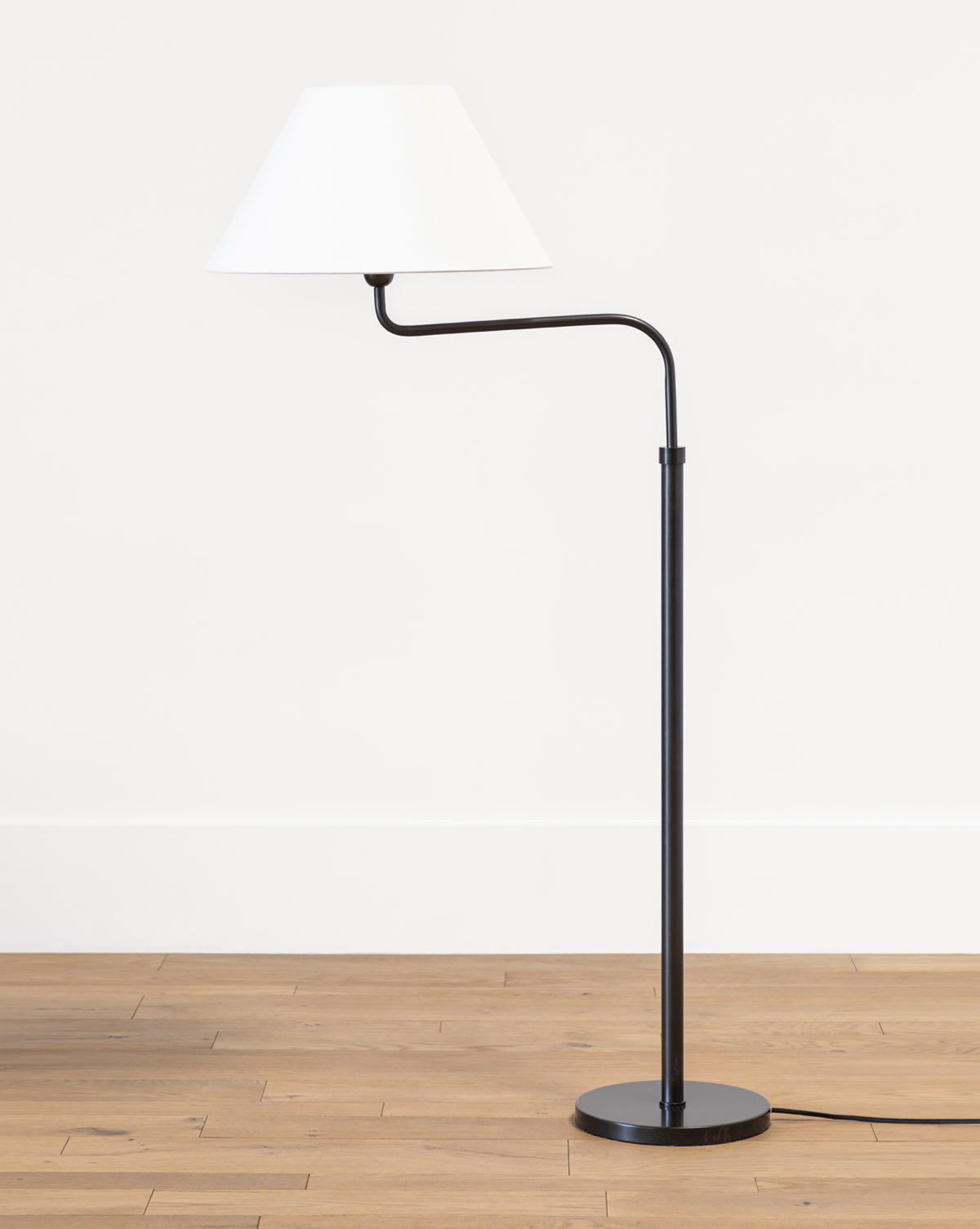 The Makerspalm Wainwright Swoop Floor Lamp features a modern black and brass slim, angled stand with a white conical shade, shown on a wooden floor against a plain white wall. McGee & Co.