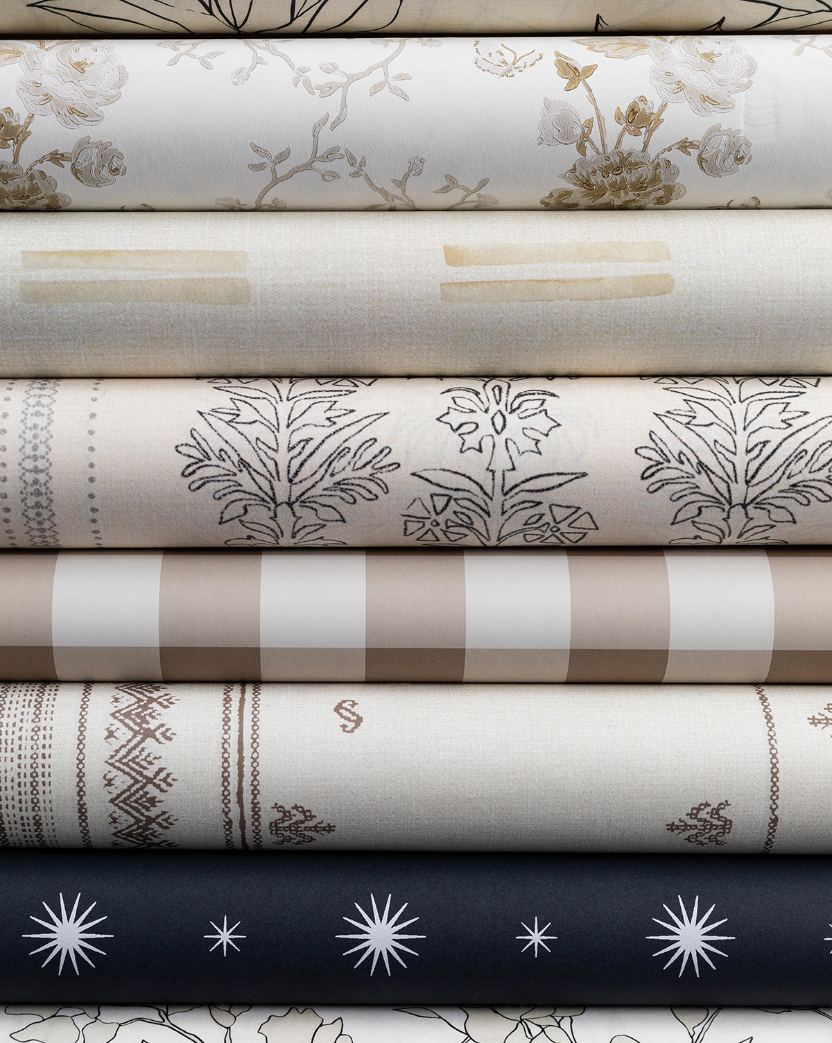 A stack of neutral-toned fabrics with florals, stripes, geometrics, and stars—echoing the Demi Floral Stripe Wallpaper by York Wallcoverings—arranged in layers on premium matte paper by McGee & Co.