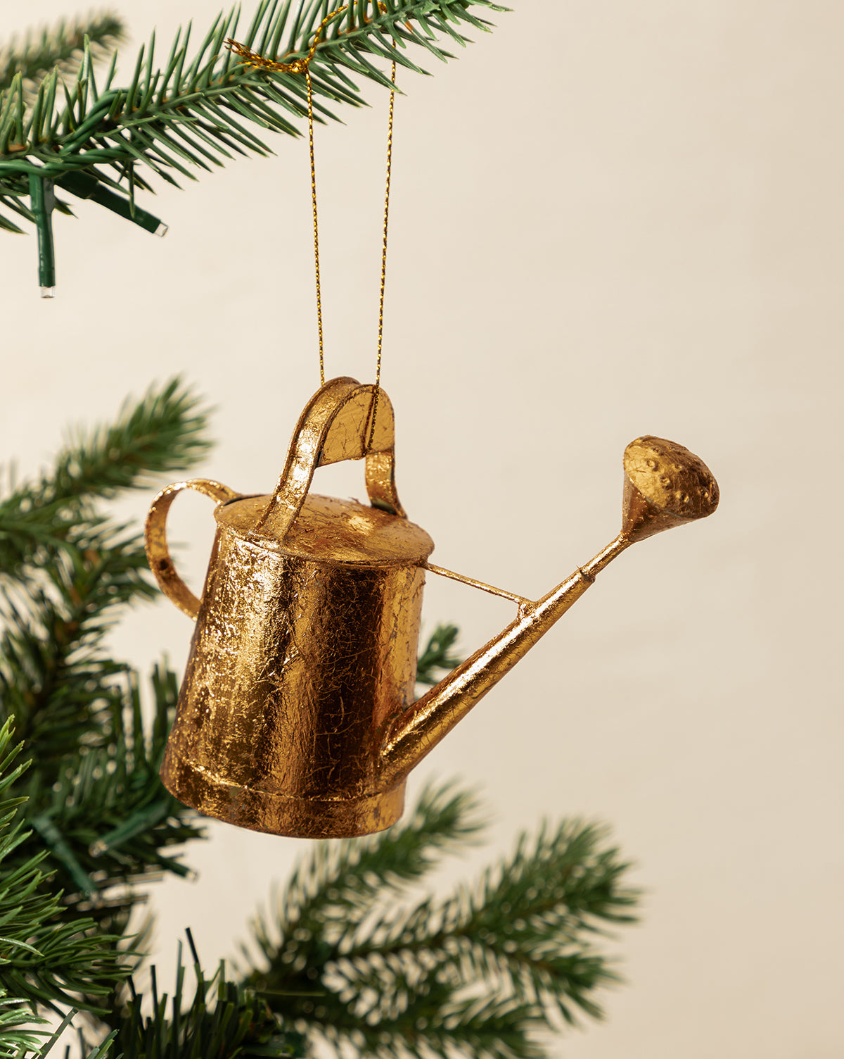 The Watering Can Ornament with a copper finish hangs from a string on a green Christmas tree branch, bringing festive charm to the holiday decor, by McGee & Co.