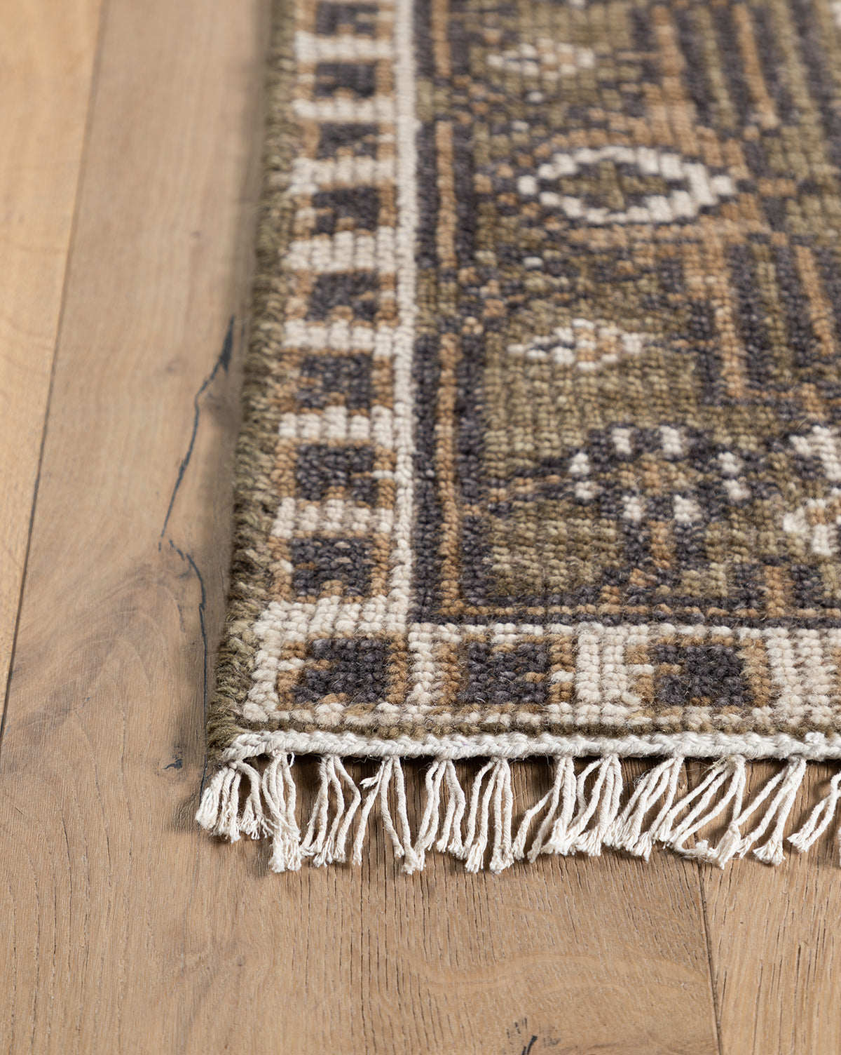 A close-up of the Wayland Hand-Knotted Wool Rug shows its decorative border and white fringe on a light wood floor. The artisan wool rug features earthy tones of brown, beige, and olive, by McGee & Co.