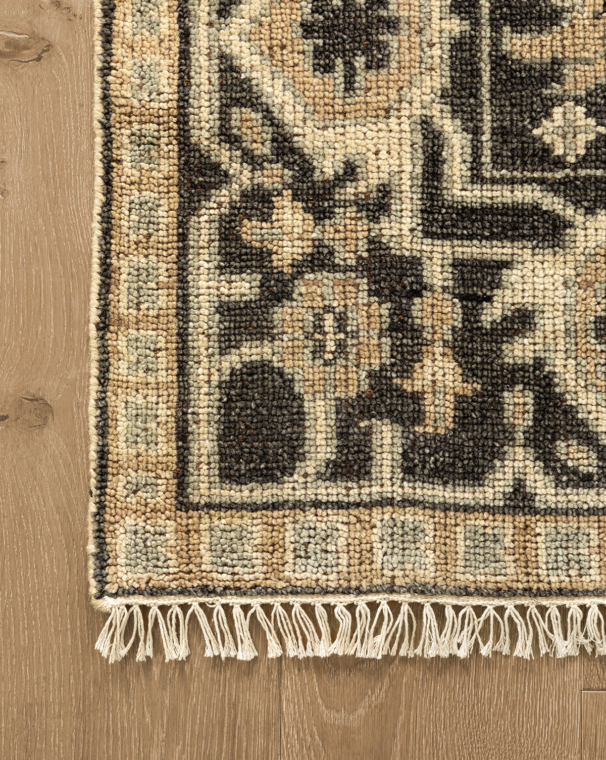A close-up of the corner of a Wayland Hand-Knotted Wool Rug highlights its beige, black, and brown floral pattern with fringe, displayed on a light wood floor by McGee & Co.