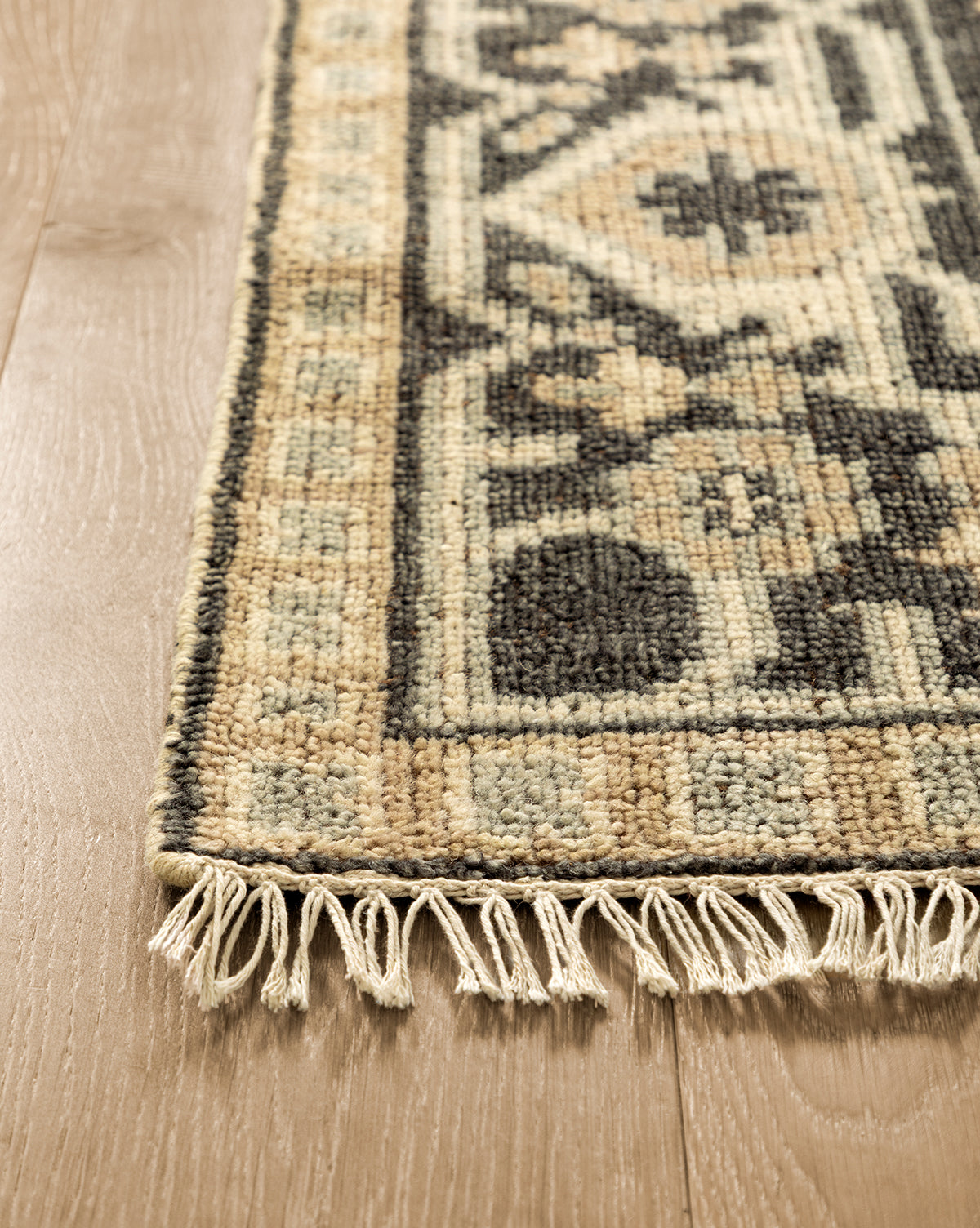 A close-up of the Wayland Hand-Knotted Wool Rug, showing beige, black, and cream patterns with fringe along the edge, laid on a light wooden floor, by McGee & Co.