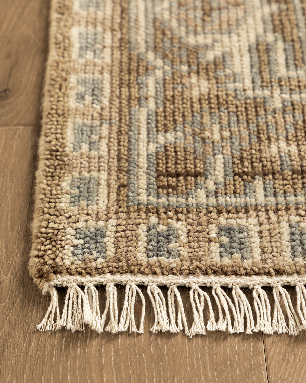 A close-up of the corner of the Wayland Hand-Knotted Wool Rug in beige, brown, and blue with knotted fringe on a wooden floor—highlighting its artisan craftsmanship, by McGee & Co.