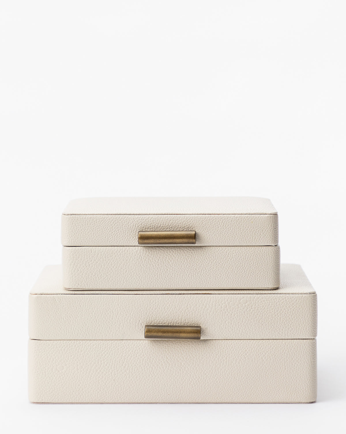 Two stacked Cream Shagreen Boxes, featuring a cream-colored textured surface and brass handles, serve as elegant decorative storage, displayed against a plain white background.
