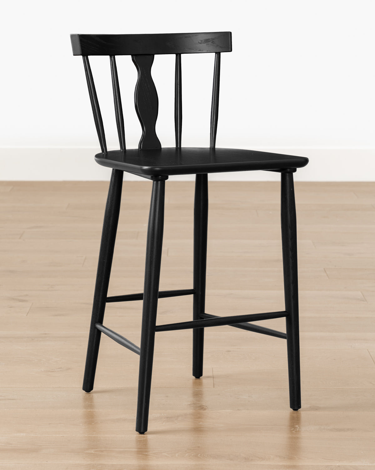 The Whittaker Counter Stool by YANG CHENG WOODEN INDUSTRIES INTERNATIONAL (VIETNAM) CO LTD features a black wood finish with a curved spindle back, three vertical spindles, and a decorative center detail, shown on a light floor against a white wall. McGee & Co.