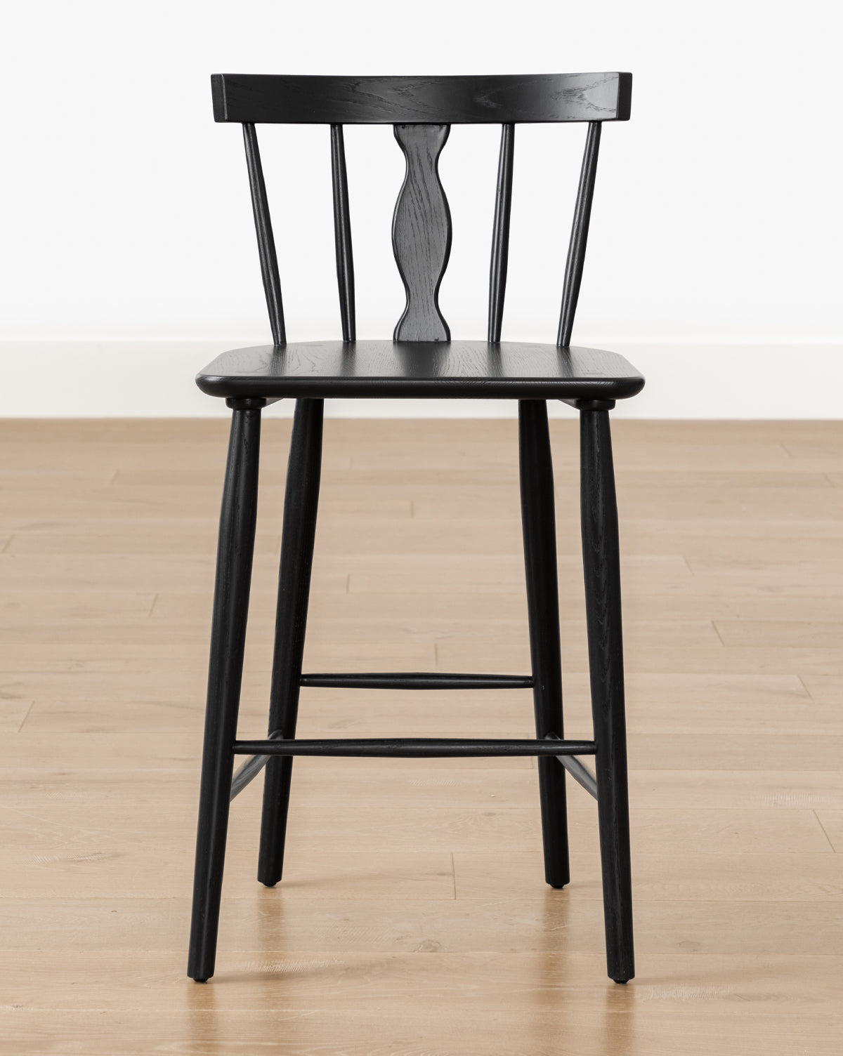 A black Whittaker Counter Stool by YANG CHENG WOODEN INDUSTRIES INTERNATIONAL (VIETNAM) CO LTD, featuring a spindle back and decorative center, crafted from solid oak, sits on a light wood floor against a plain white wall, McGee & Co.