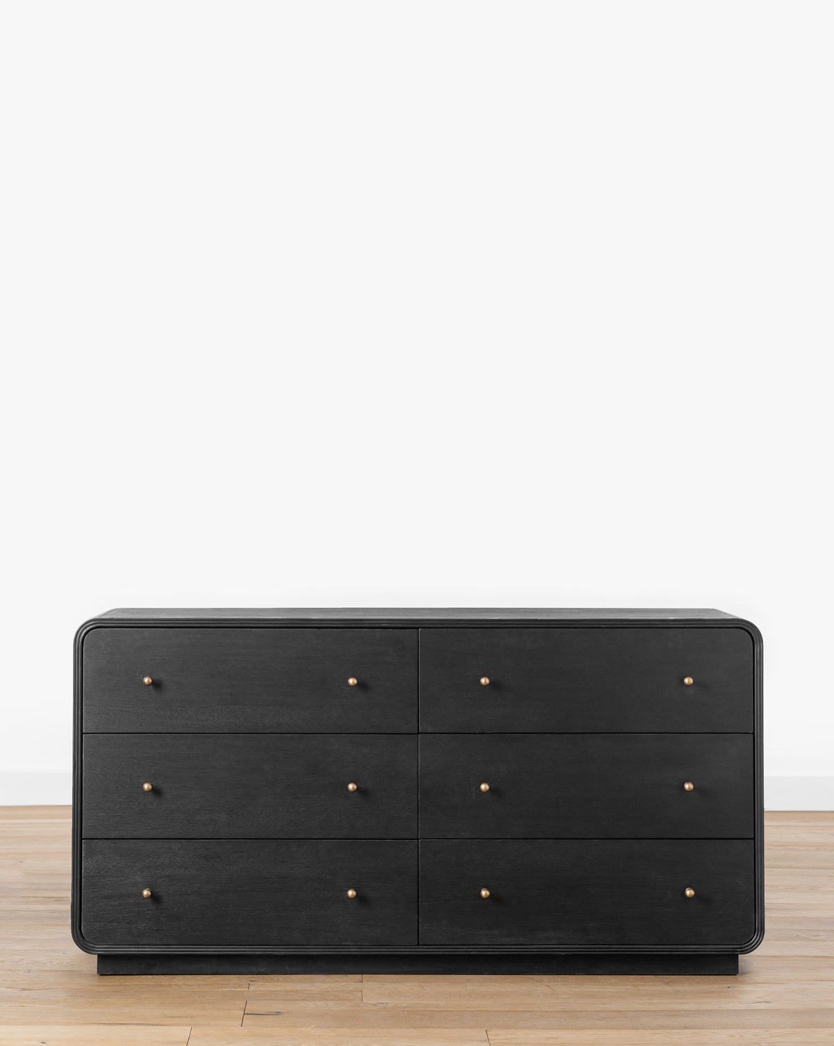 The Wilkins Dresser is a modern black acacia wood dresser with six soft-close drawers, each with a small round gold knob, shown on a light wood floor against a plain white background, by McGee & Co.