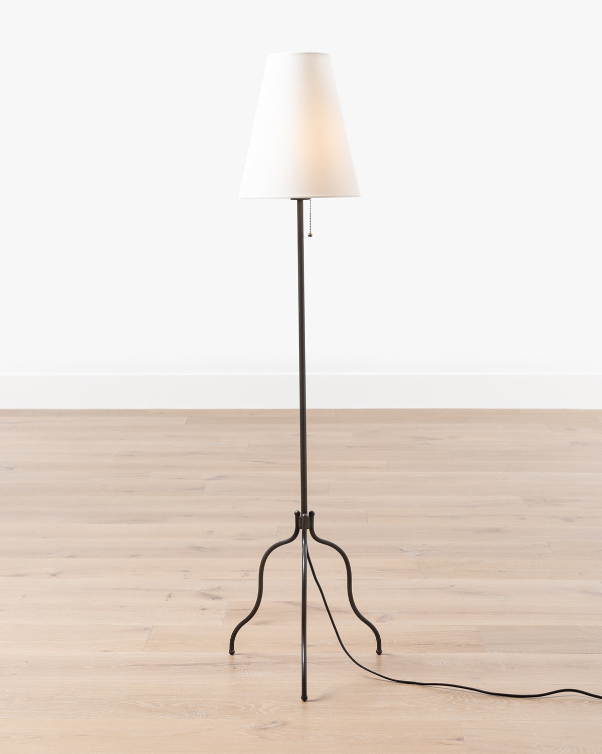 The Windham Floor Lamp stands gracefully on a light wood floor against a white wall, featuring a white linen shade perched on a slender matte black stand with three curved legs. Its power cord extends to the right, by McGee & Co.