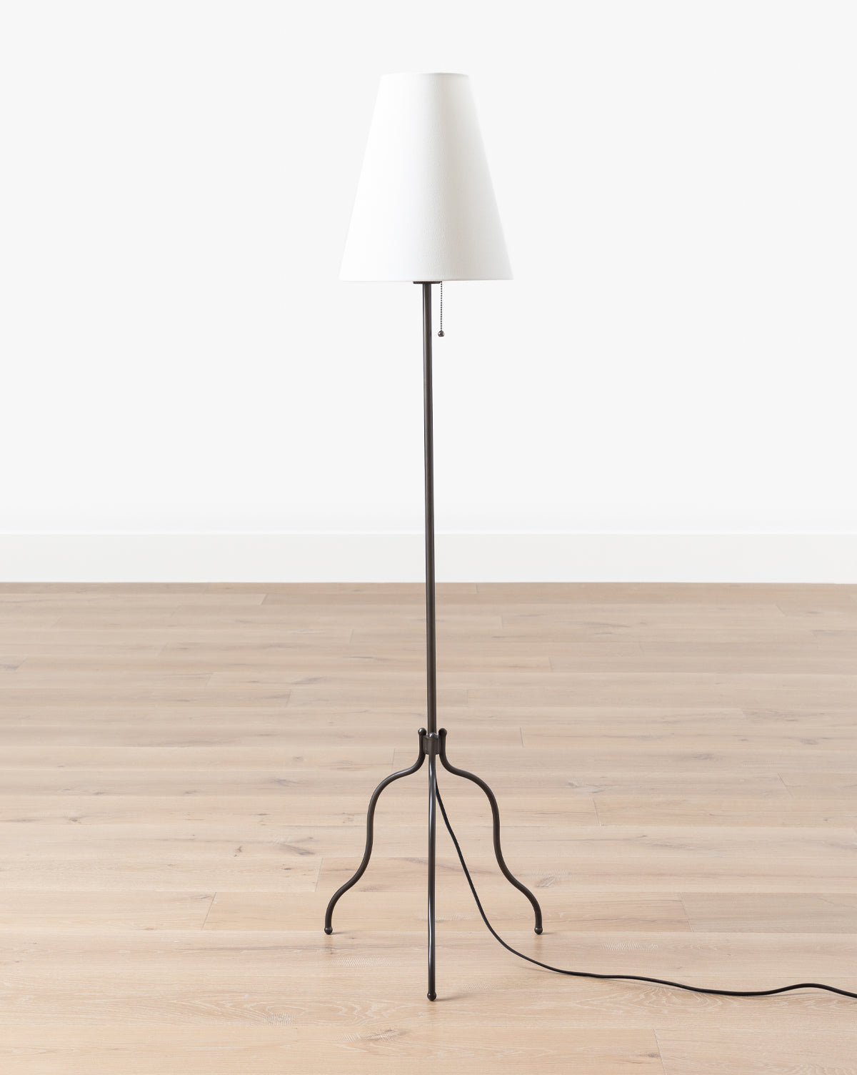 The Makerspalm Windham Floor Lamp features a minimalist design with a white linen shade and slender matte black tripod base, standing on a light wood floor against a plain white wall. Its power cord extends across the floor. McGee & Co.