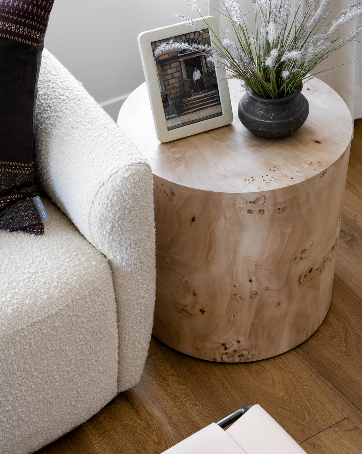 A round, light wood side table displays a Shagreen Picture Frame and a dark vase with green foliage. Part of a textured white sofa and wood floor are also visible.