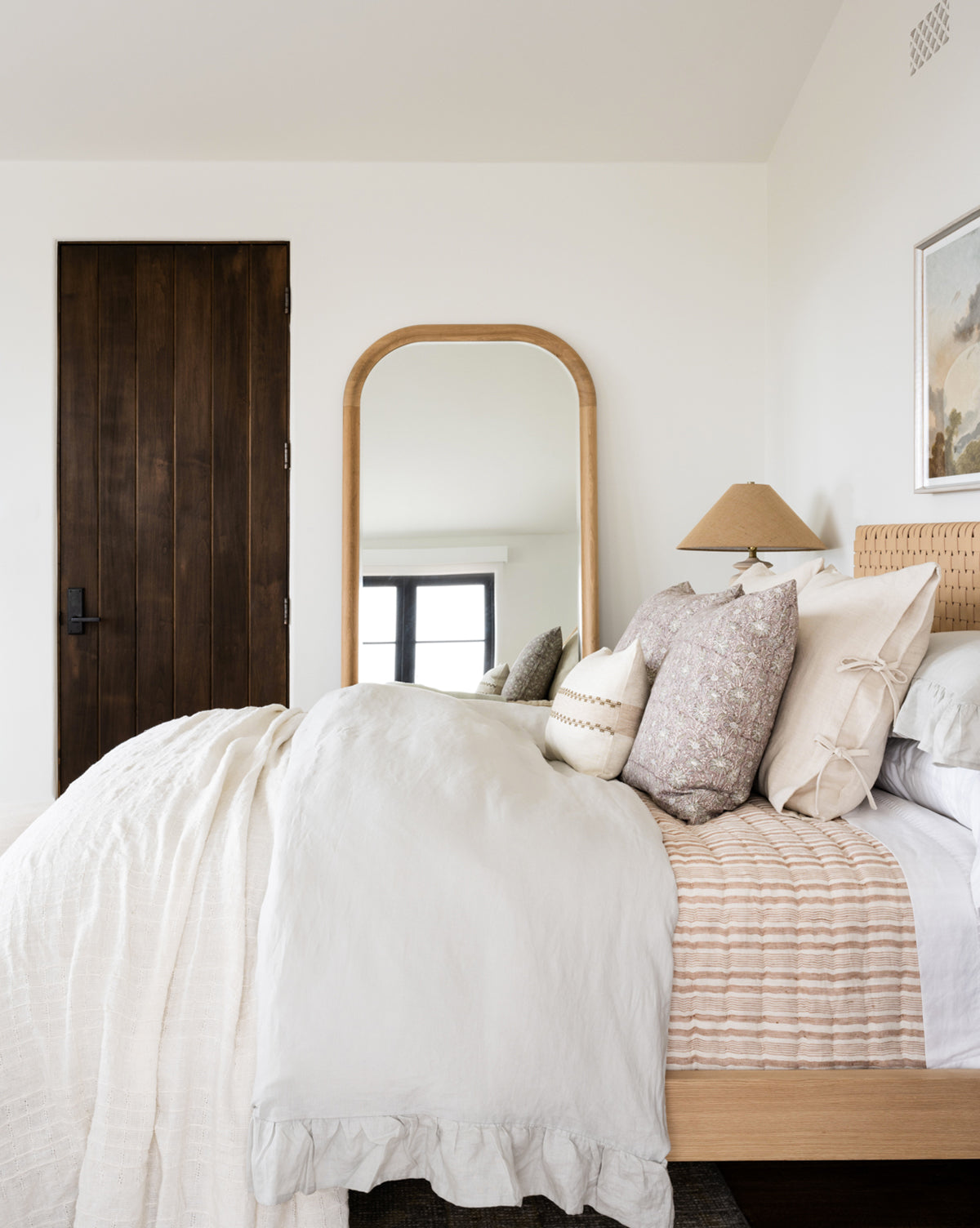 A cozy bedroom with neutral tones features a wooden bed dressed in the Ruffle Linen Duvet Cover, layered pillows, a cane headboard, framed art, a large arched mirror, and a dark wood door.
