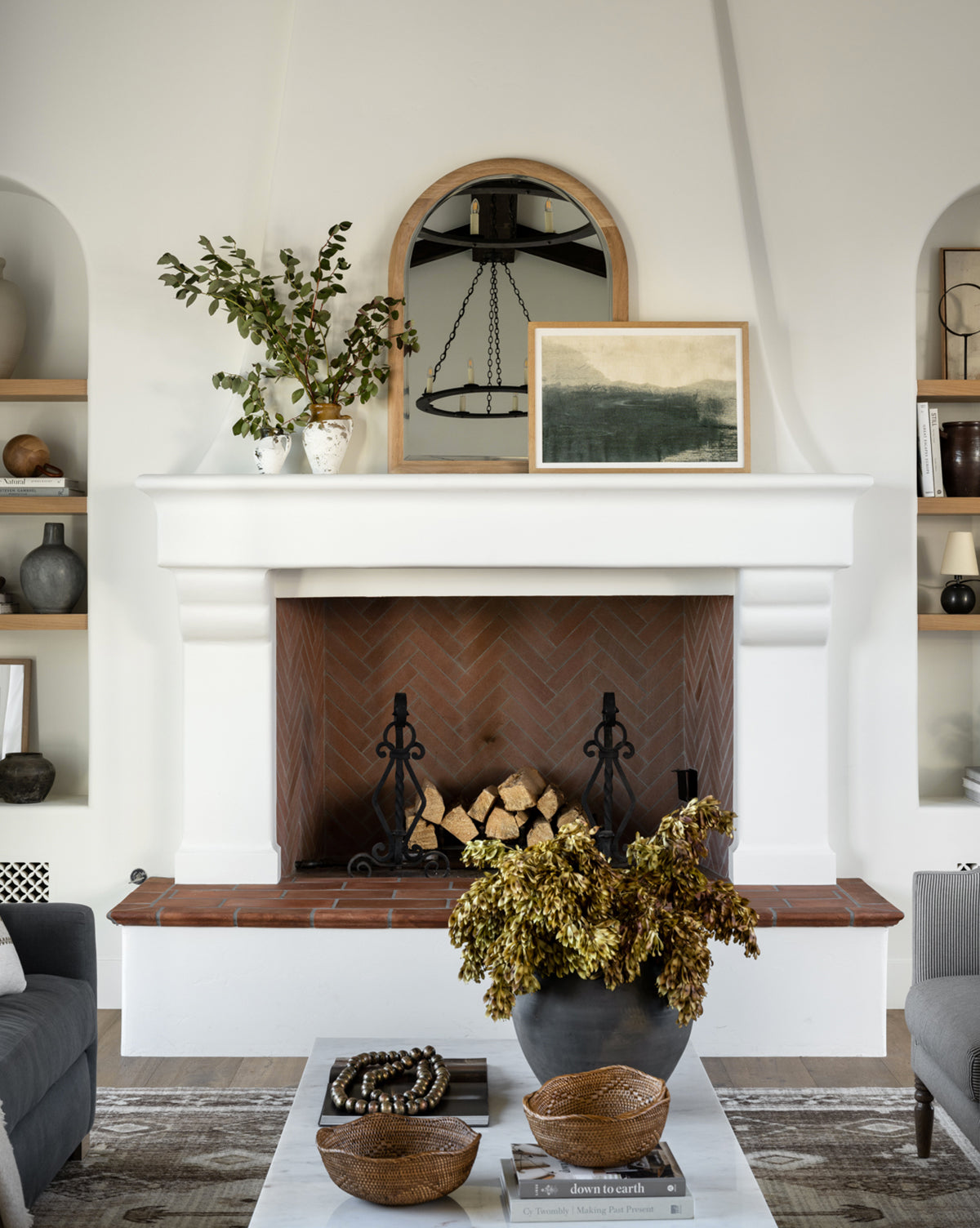 A stylish living room features a white fireplace with firewood, a Reta Arched Wall Mirror above the mantel, framed art and vases with greenery. Natural oak shelves hold books and decor; the coffee table displays bowls and a floral bouquet by McGee & Co.