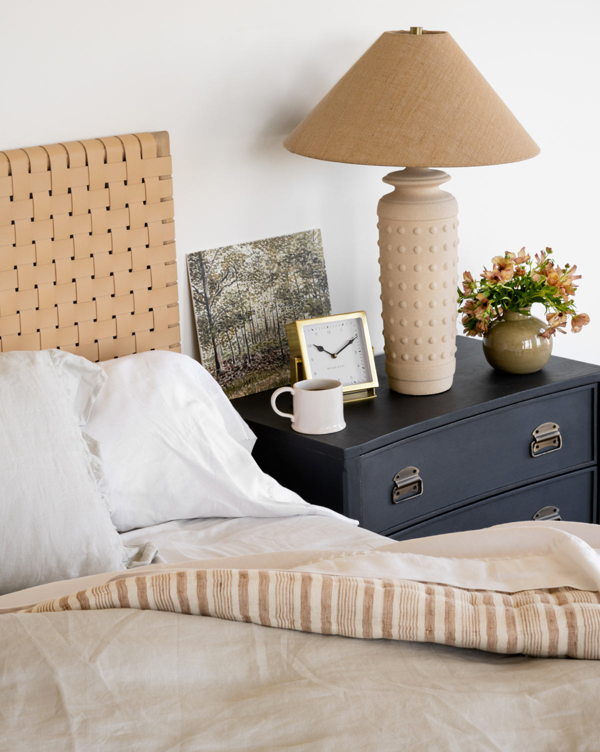 A cozy bedroom showcases an unmade bed with beige and white bedding, a woven headboard, and a dark nightstand displaying a ceramic lamp, the Posey Table Clock, a mug, fresh flowers in a vase, and a framed landscape photo.