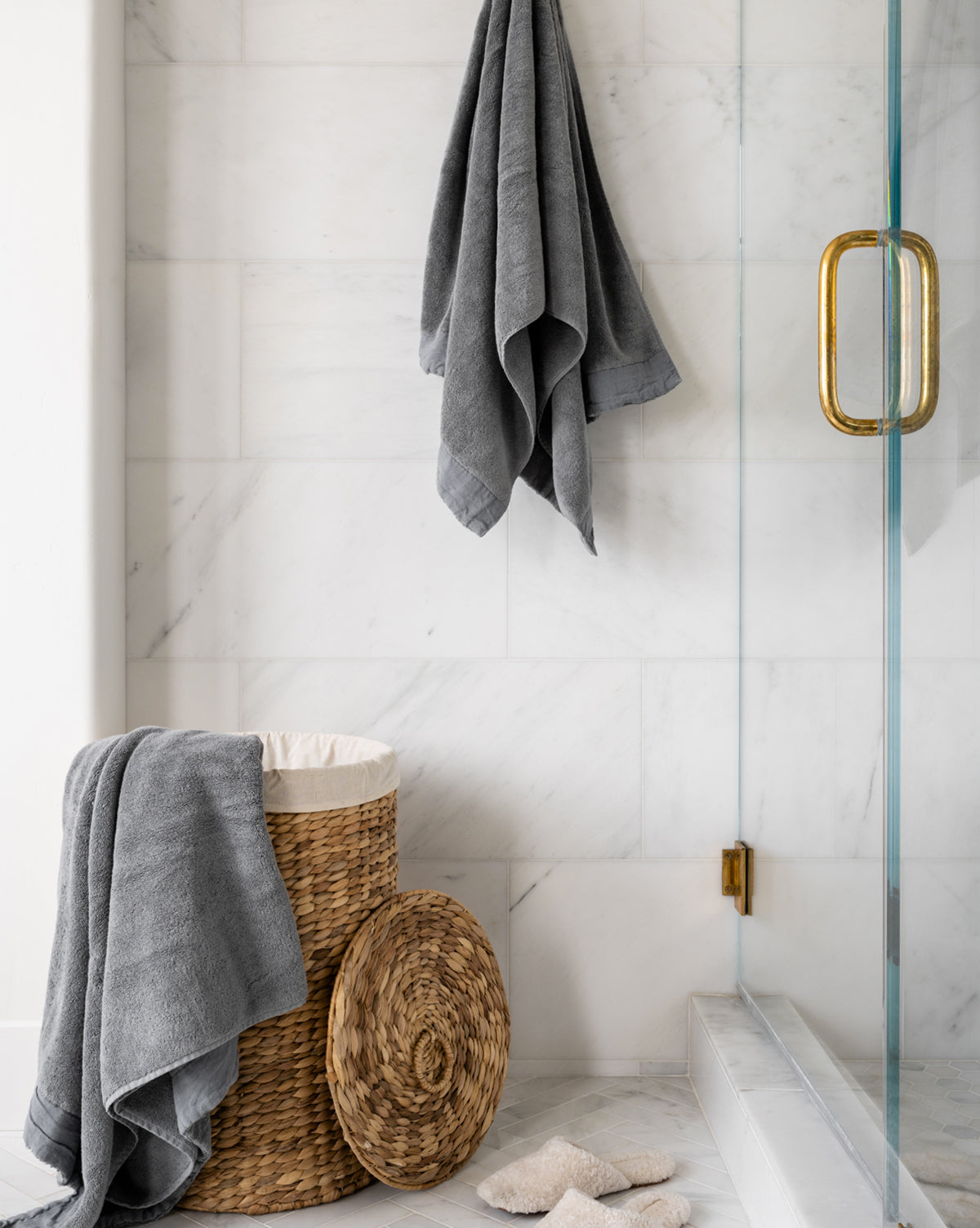 A modern bathroom with white marble walls, a glass shower door with a gold handle, and the Round Water Hyacinth Basket adding warmth. A gray towel drapes over the laundry basket, with slippers on the floor and extra gray towels hanging nearby.