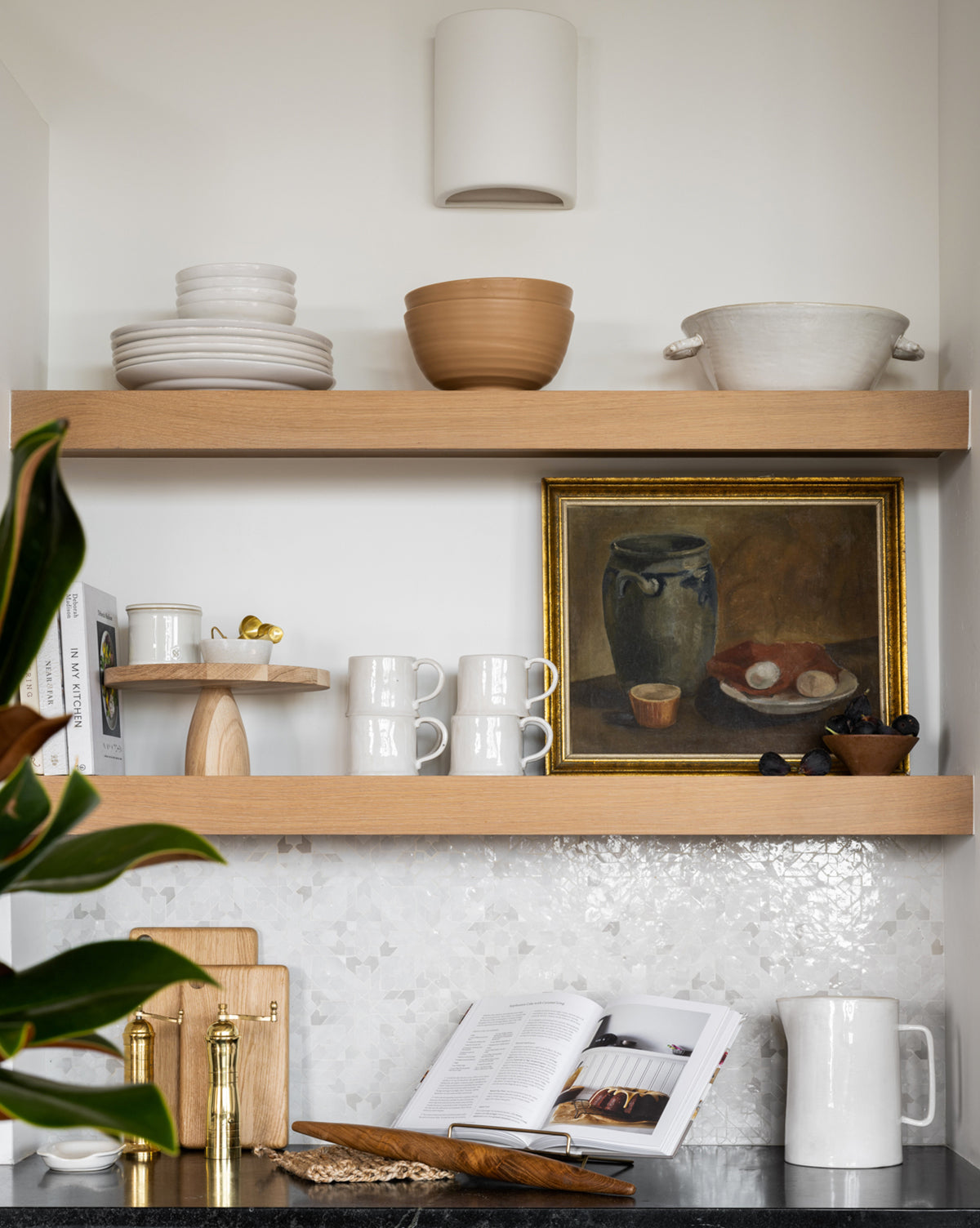 Open wooden shelves display dishes, mugs, and bowls with a framed painting above. On the marble countertop sits an open book near a plant, utensils, white pitcher, and the Wooden Cake Stand for stylish home decor, by McGee & Co.