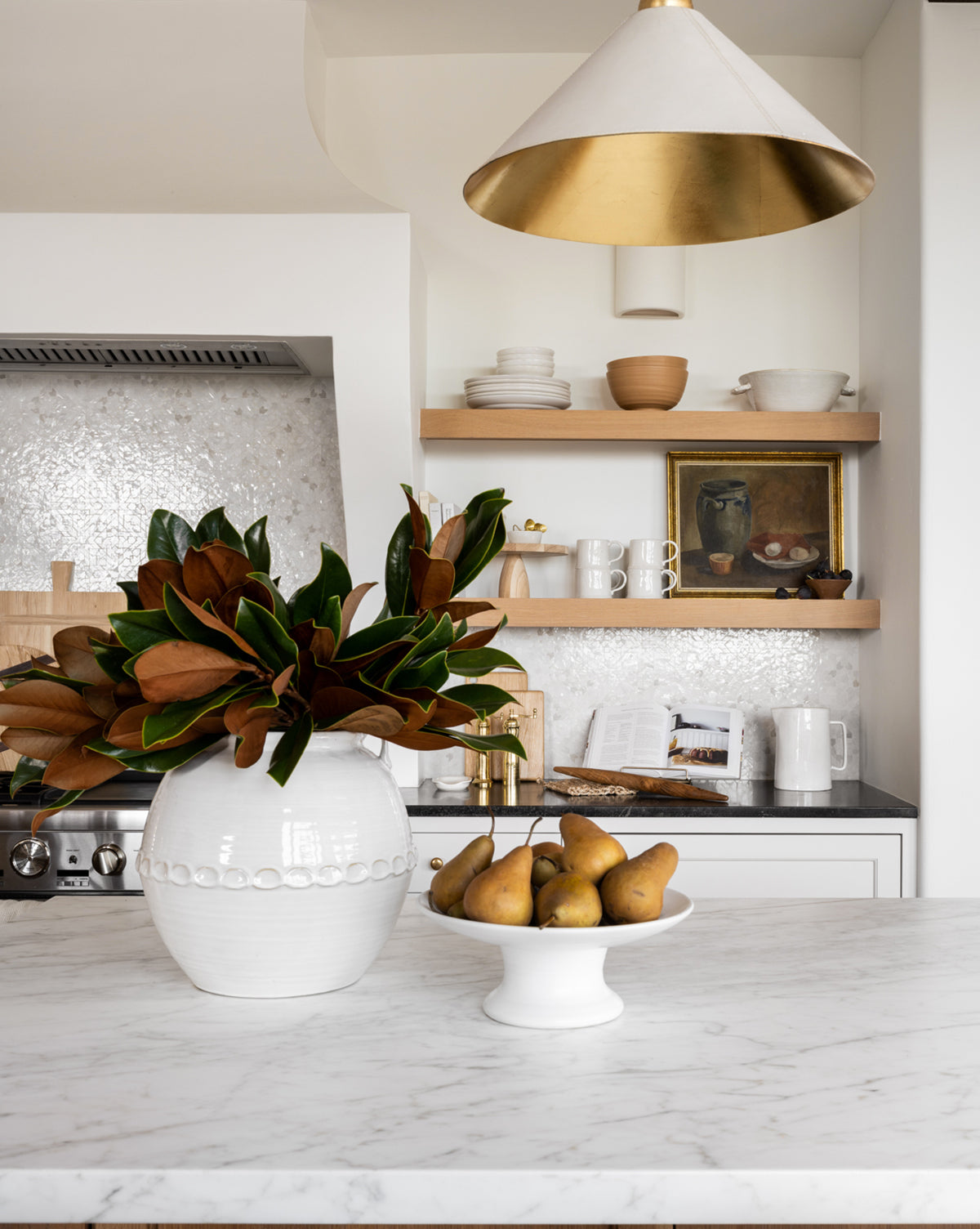 A modern kitchen with a marble countertop, a Rounded White Vase filled with leafy branches, a bowl of pears, open wooden shelves displaying dishes, a gold and white pendant light, and a metallic tile backsplash.