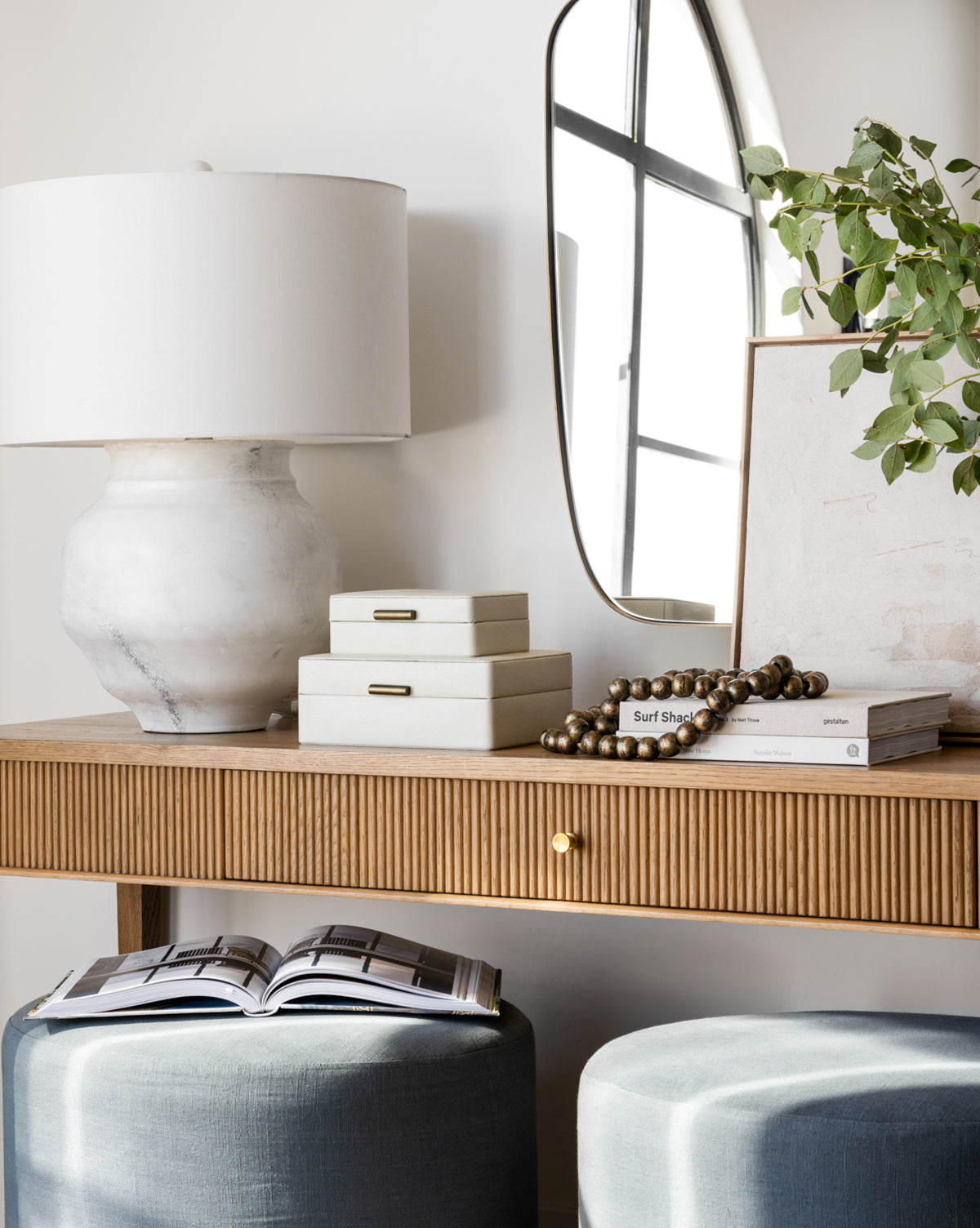 A modern wooden console table is styled with a white lamp, books, bead garland, leafy plant, the Cream Shagreen Box, and an arched mirror. Below are two blue ottomans and an open magazine.