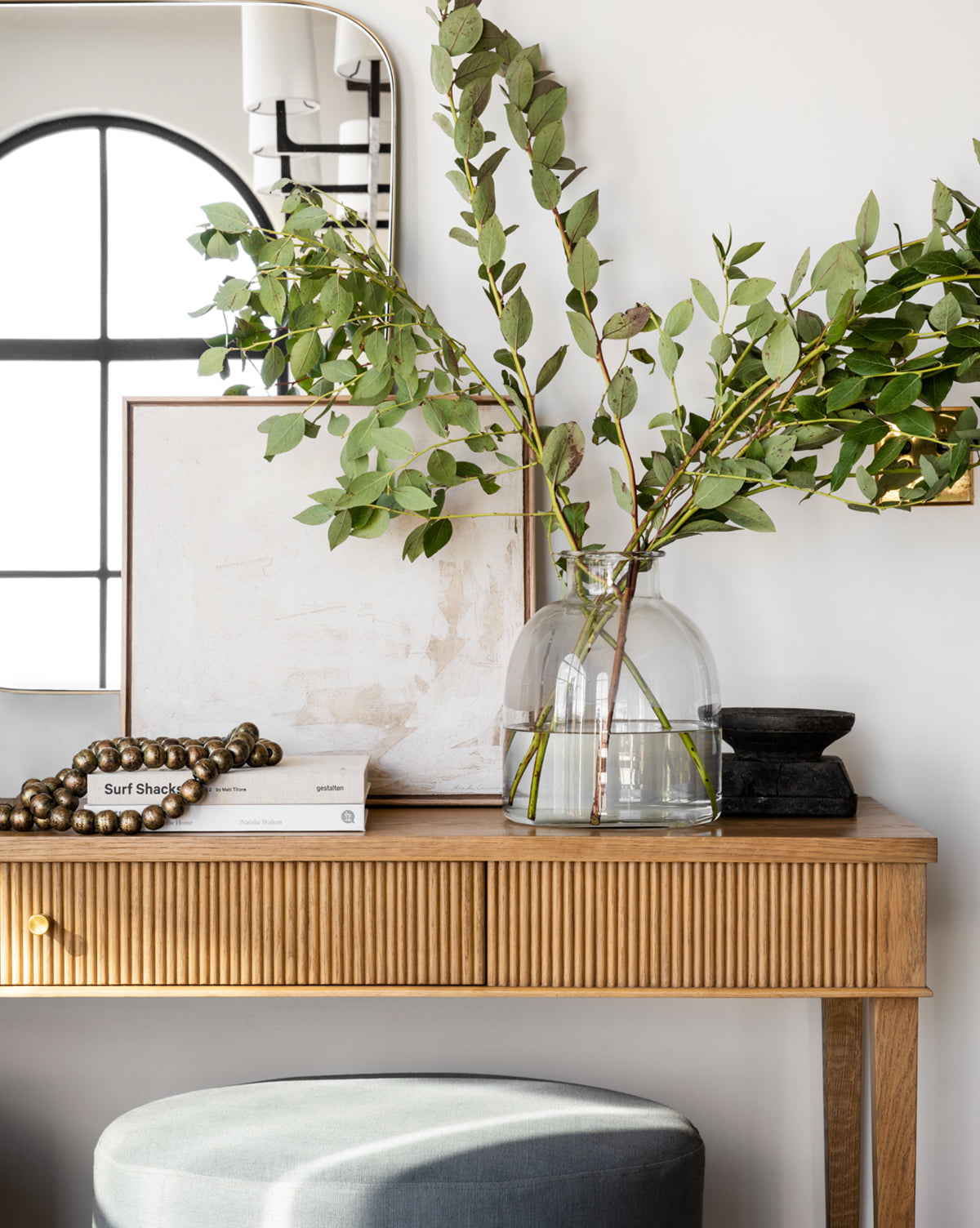 A Kelby Glass Vase filled with green branches rests on a wooden console table with books, a bead garland, and black accents. Behind, theres framed art and a mirror.