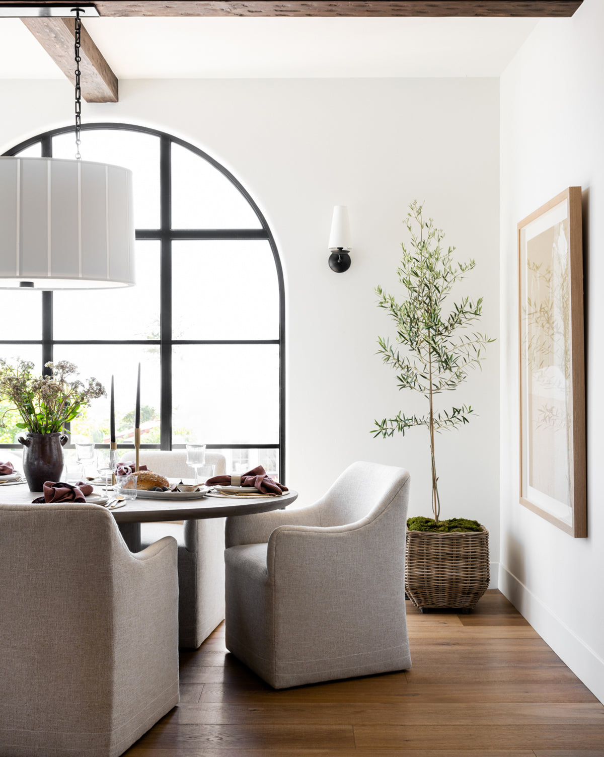 Bright dining area with a round table and four Elton Dining Chairs in light linen, large arched window, potted plant, framed wall art, wood flooring; table is set with plates and a floral centerpiece—ideal for stylish dining by McGee & Co.