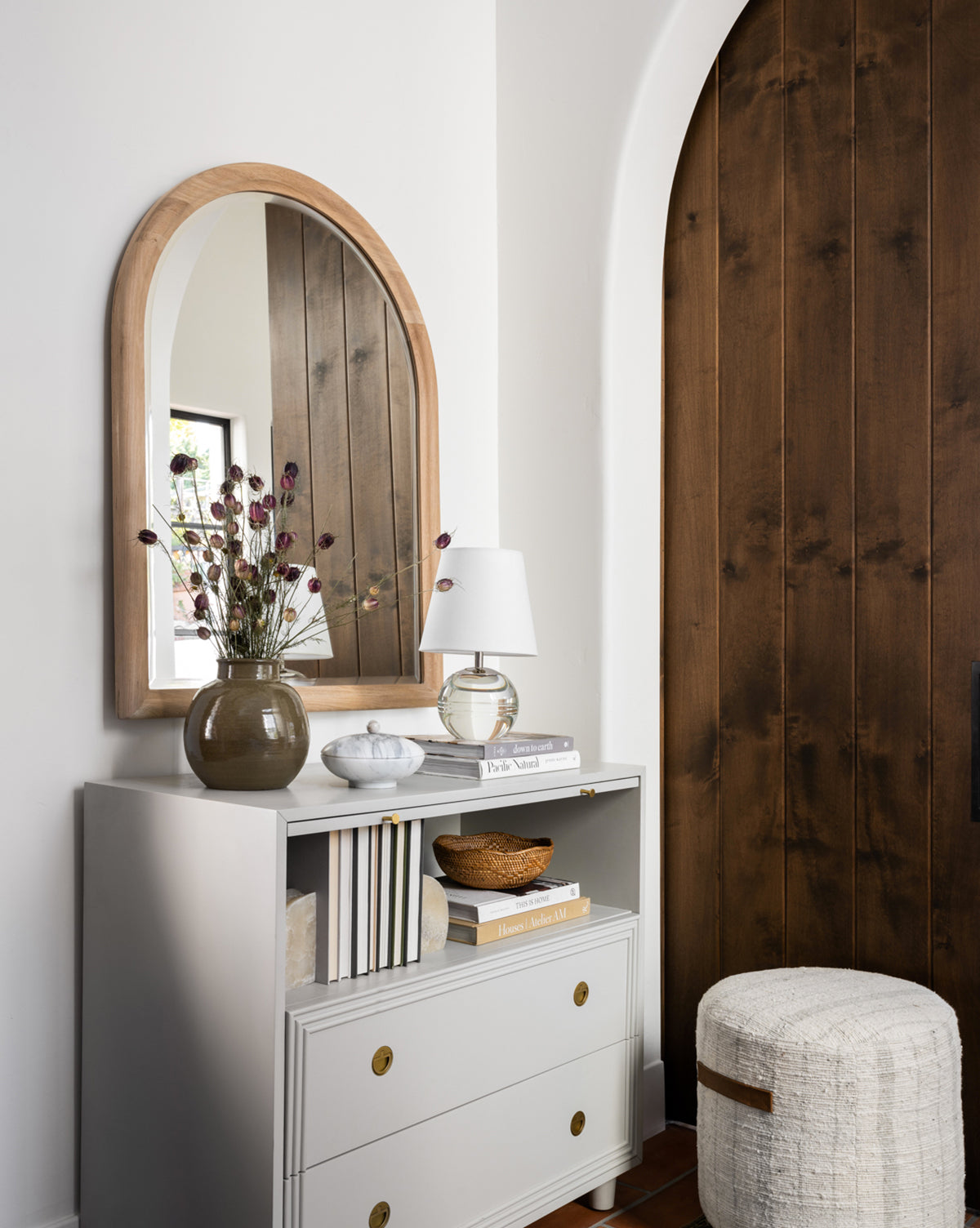A white dresser with gold handles holds books, a small white lamp, and a brown vase with dried flowers. Above it hangs the Reta Arched Wall Mirror reflecting a window. Nearby are a wooden arched door and a textured round ottoman by McGee & Co.