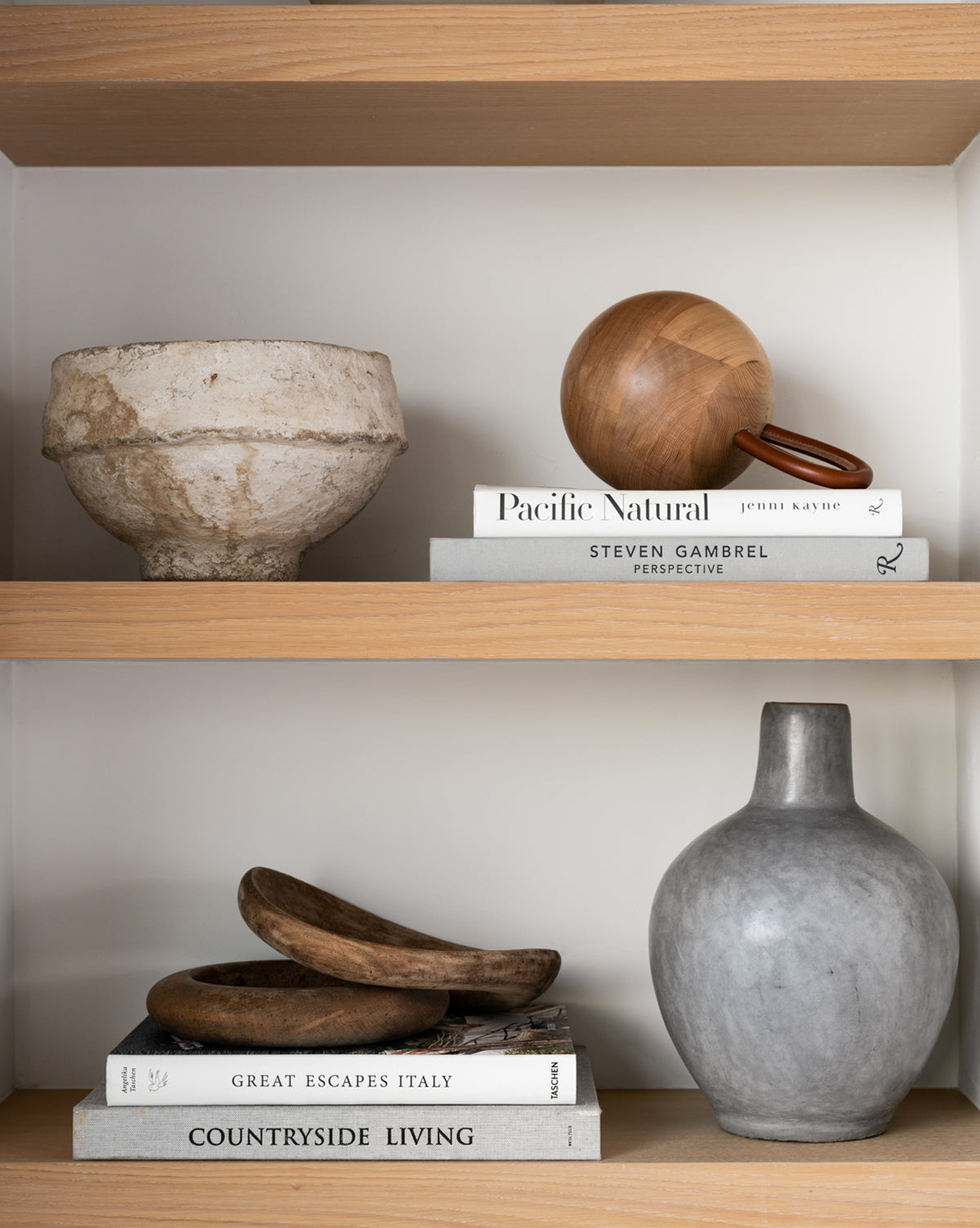 Two wooden shelves showcase a rough ceramic bowl, wooden sphere, art books, trays, and a matte gray vase—bringing the minimalist elegance of stunning hotels and Great Escapes Italy into your home.