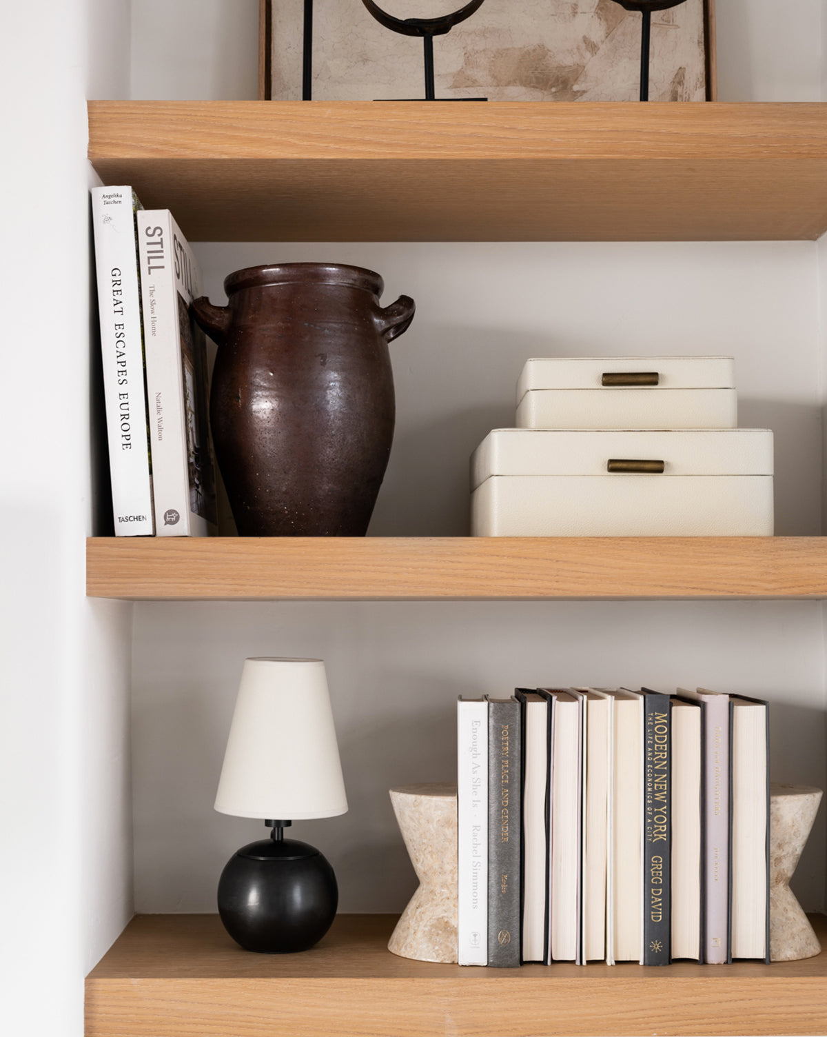 Three wooden shelves display decor: the top shelf holds books, a brown ceramic vase, and white boxes; the middle has white boxes and books; the bottom features the Tiny Terri Accent Lamp with a natural paper shade, books, and a textured decorative piece by McGee & Co.