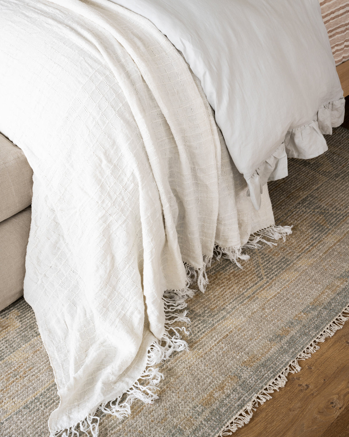 A close-up shows a neatly made bed with a white textured blanket and fringed ends cascading onto the Roslin Hand-Knotted Wool Rug, featuring neutral tones and a subtle geometric pattern on the wooden floor, by McGee & Co.