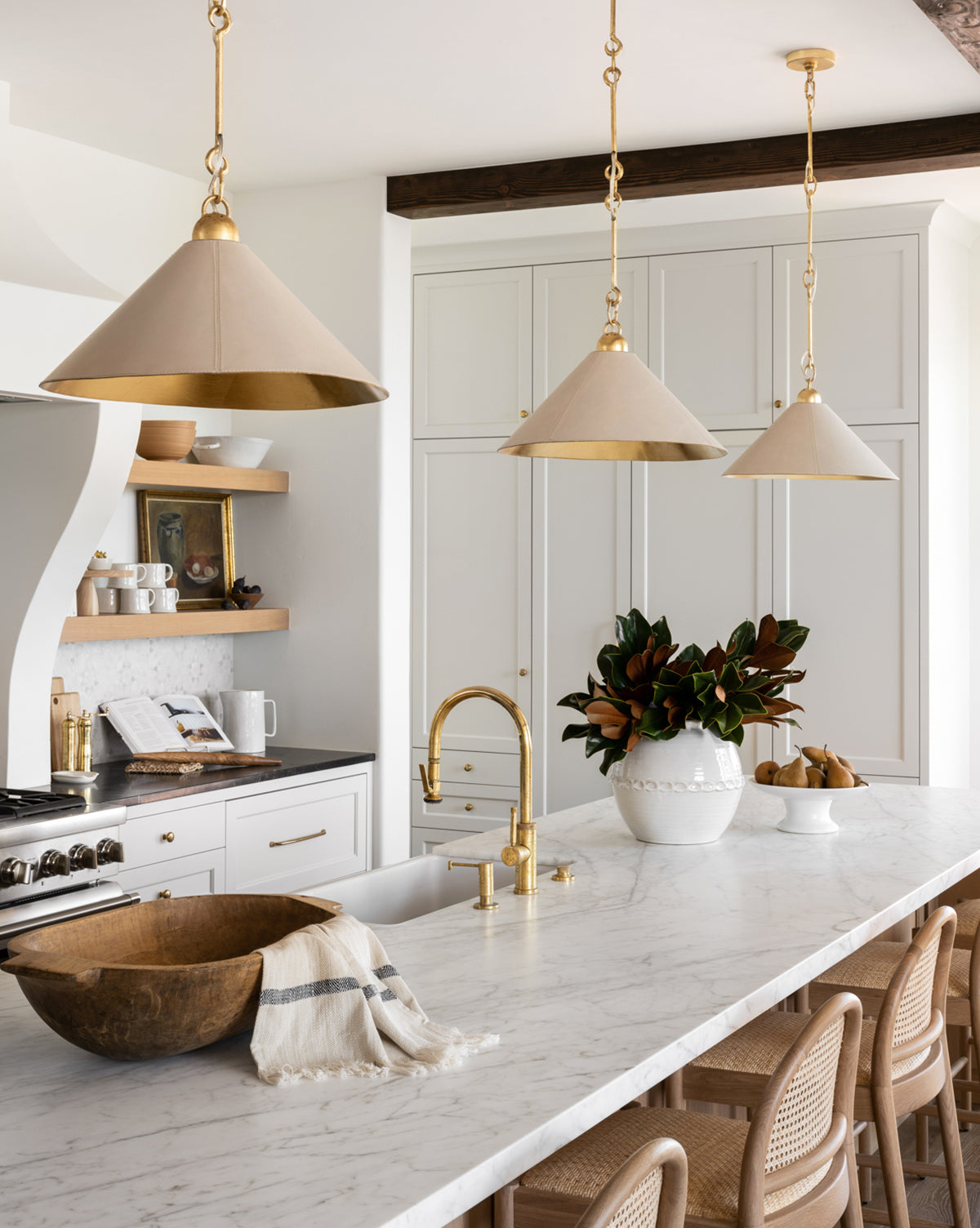 A bright kitchen features white cabinets, marble countertops, gold fixtures, beige pendant lights, woven chairs, a wooden bowl with a Fringed Striped Tea Towel, and a vase of leafy branches on the large island, by McGee & Co.