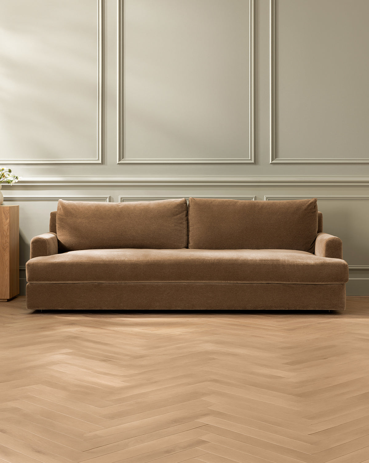The Winthrop Sofa 90, upholstered in brown with two back cushions, sits in a minimal room with light gray paneled walls and light wood herringbone flooring, by McGee & Co. A small wooden side table is partially visible on the left.