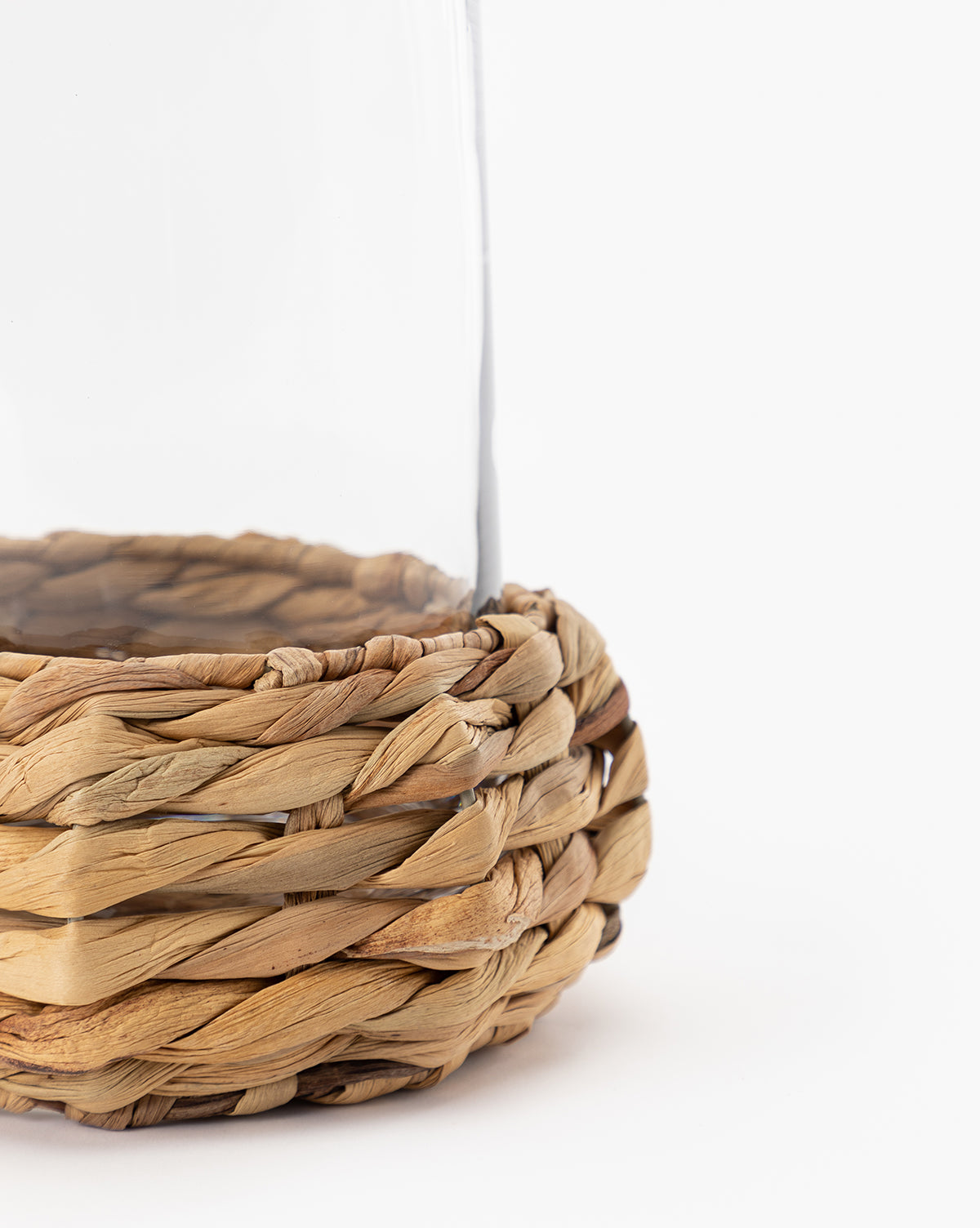 Close-up of the lower part of the Woven Hurricane, featuring a clear glass top and a natural-colored wicker base. Perfect for candle displays, this rustic accent is crafted from natural materials and set against a plain white background.