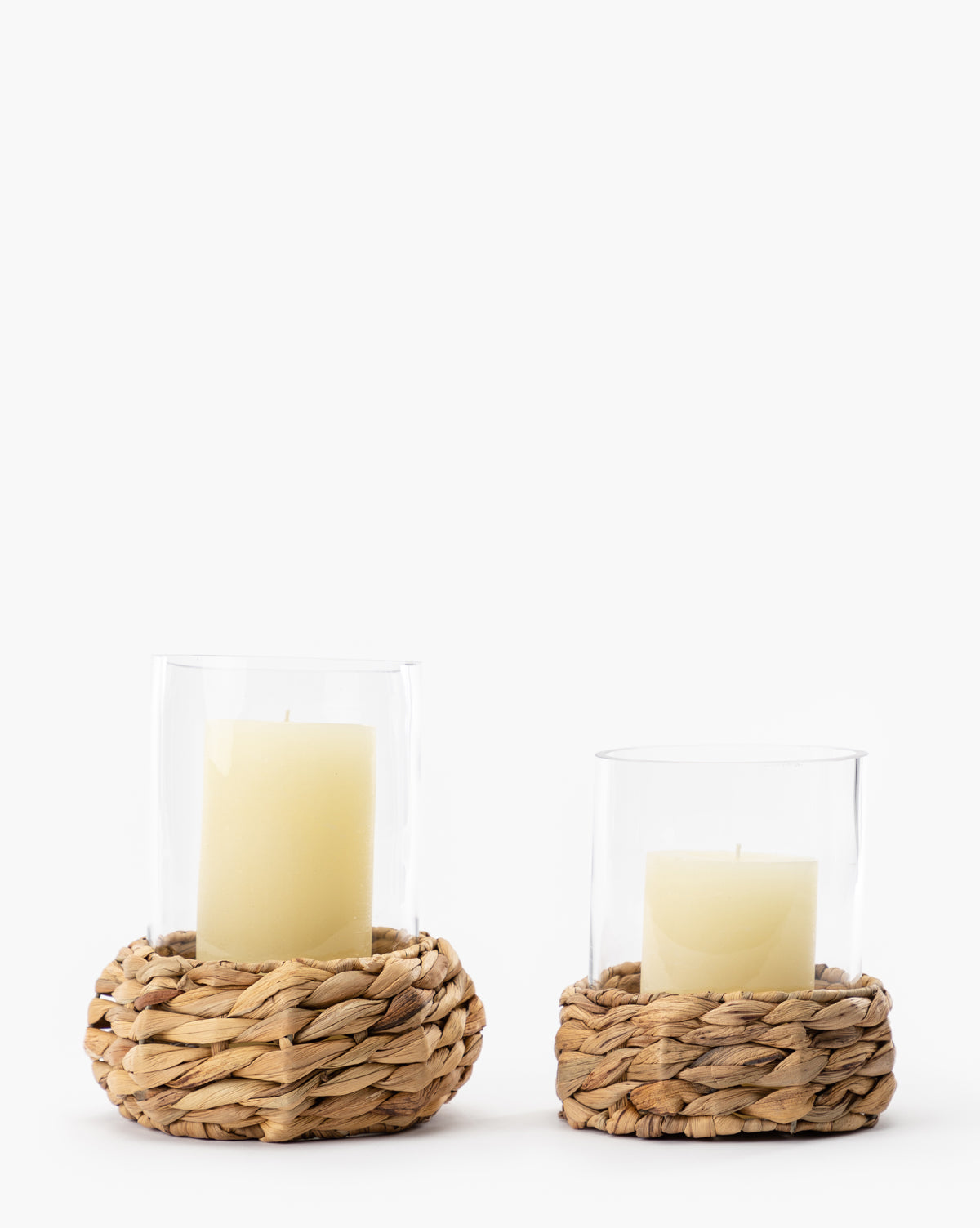 The Woven Hurricane set features two pillar candles in clear glass holders with natural-colored woven straw bases, one taller than the other, beautifully displayed against a simple white background.