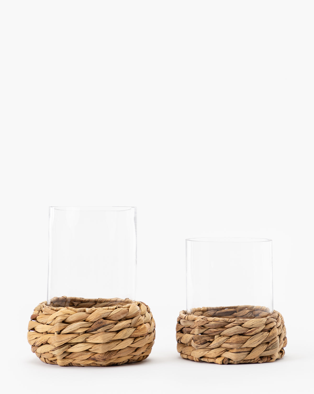 Two Woven Hurricane vases with clear glass sit in round, woven bases made from natural materials. The left vase is taller and the right one is shorter. Both are displayed against a plain white background.