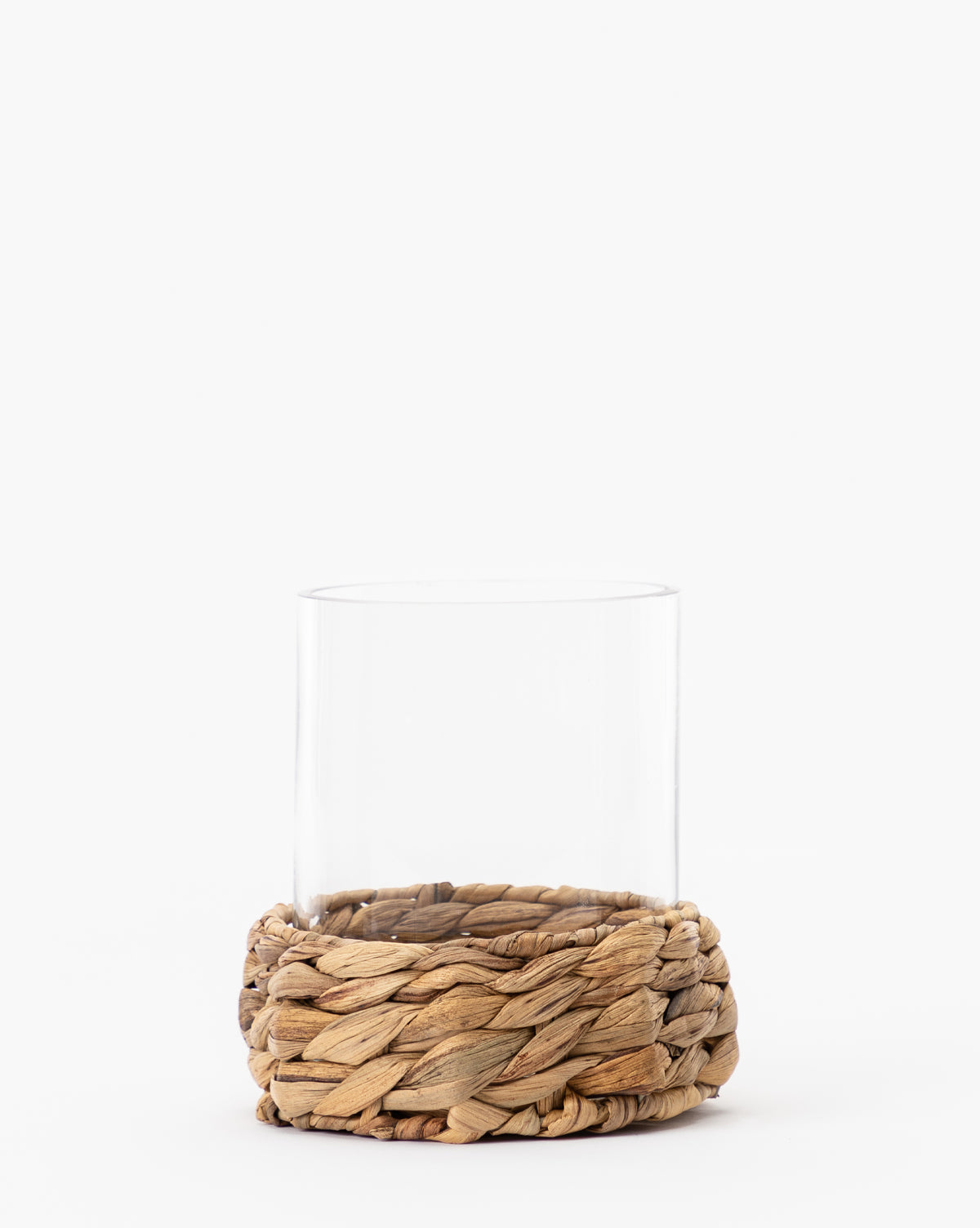The Woven Hurricane is a clear glass tumbler featuring a thick, woven natural fiber sleeve at the base—ideal for candle displays or adding a natural accent to your space. Displayed here against a plain white background.