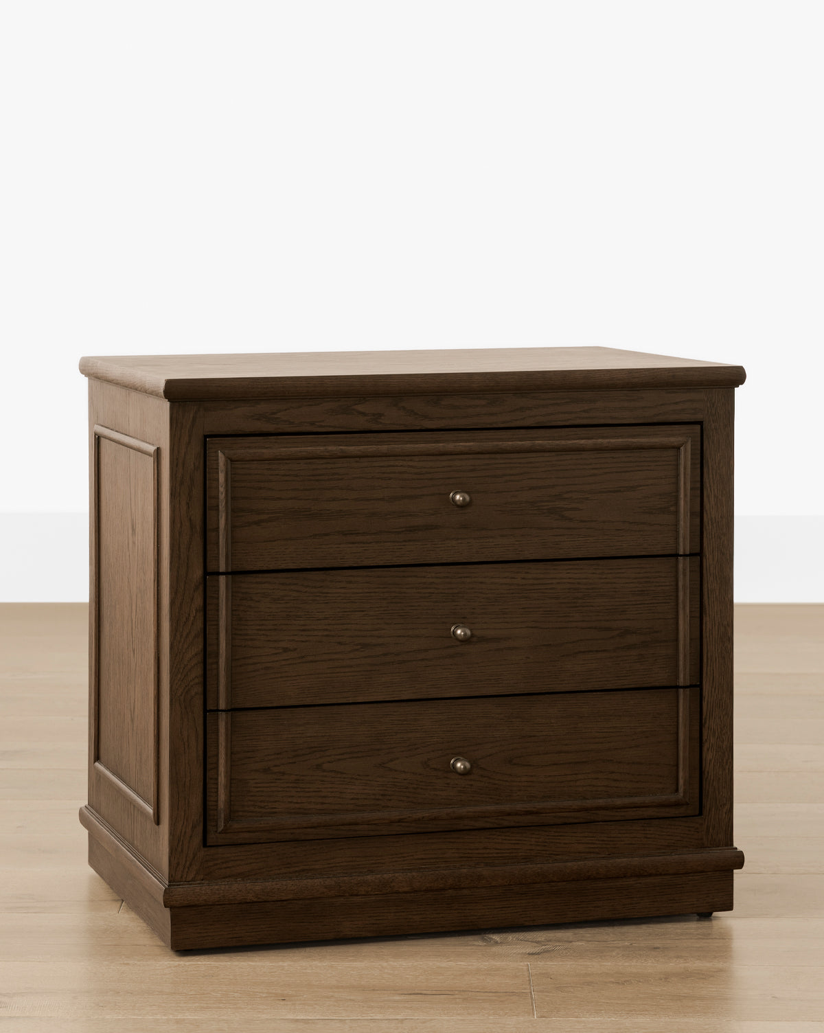 The Yorkshire Nightstand, finished in English oak, features three drawers with antique brass pulls and is displayed on a light wooden floor against a white background, by McGee & Co.