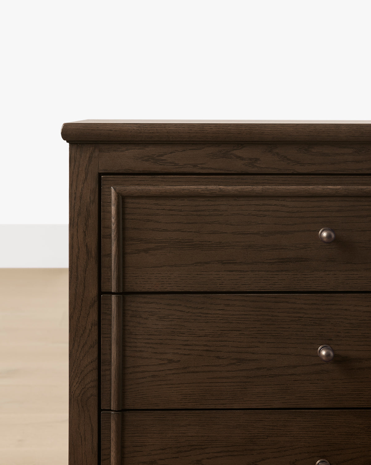 A close-up of the Yorkshire Nightstand’s top left corner reveals its English oak finish, three drawers with round antique brass pulls, and a dark wooden build set against a light floor and white wall, by McGee & Co.