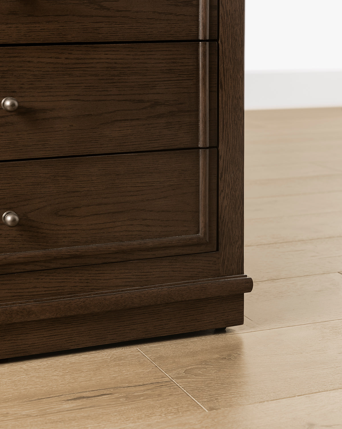 Close-up of the bottom corner of the Yorkshire Nightstand in an English oak finish with antique brass pulls, placed on a light wood floor against a white wall by McGee & Co.